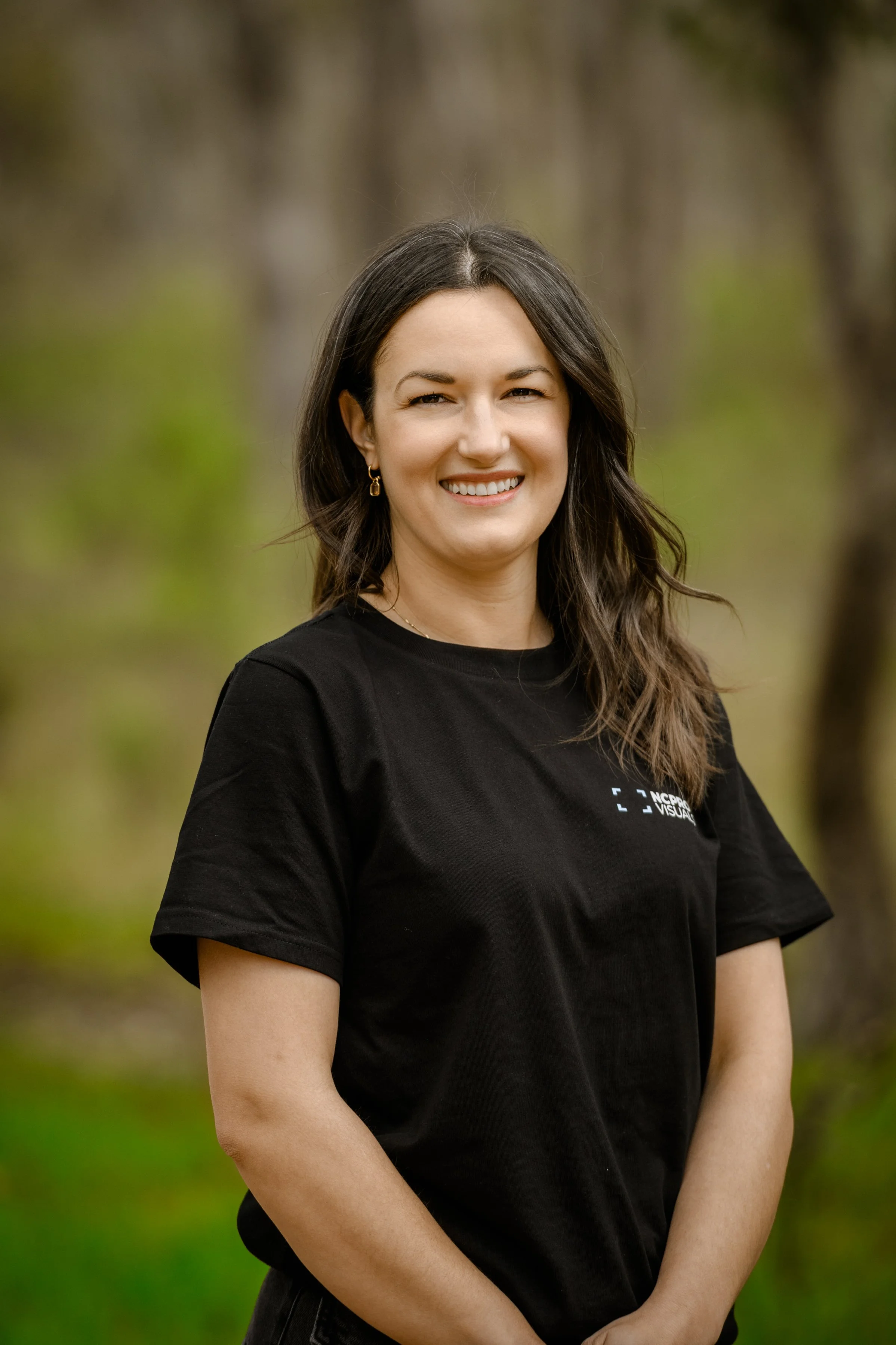 A woman with long brown hair tied back, wearing a black T-shirt with a logo, standing outdoors in a wooded area.