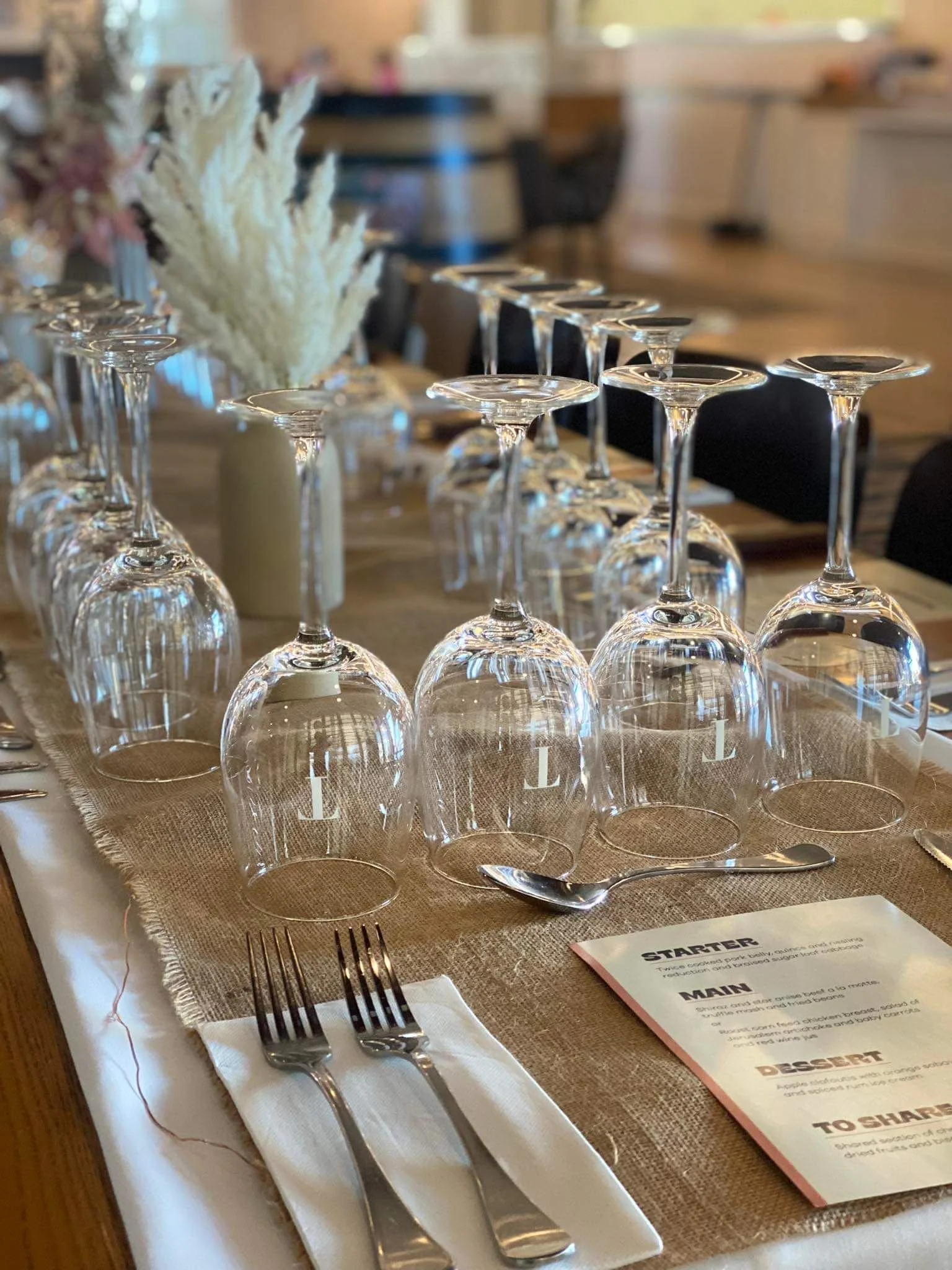 Table set with upside-down wine glasses, silverware, a menu, a spoon, candles, and decorative flowers in the background.