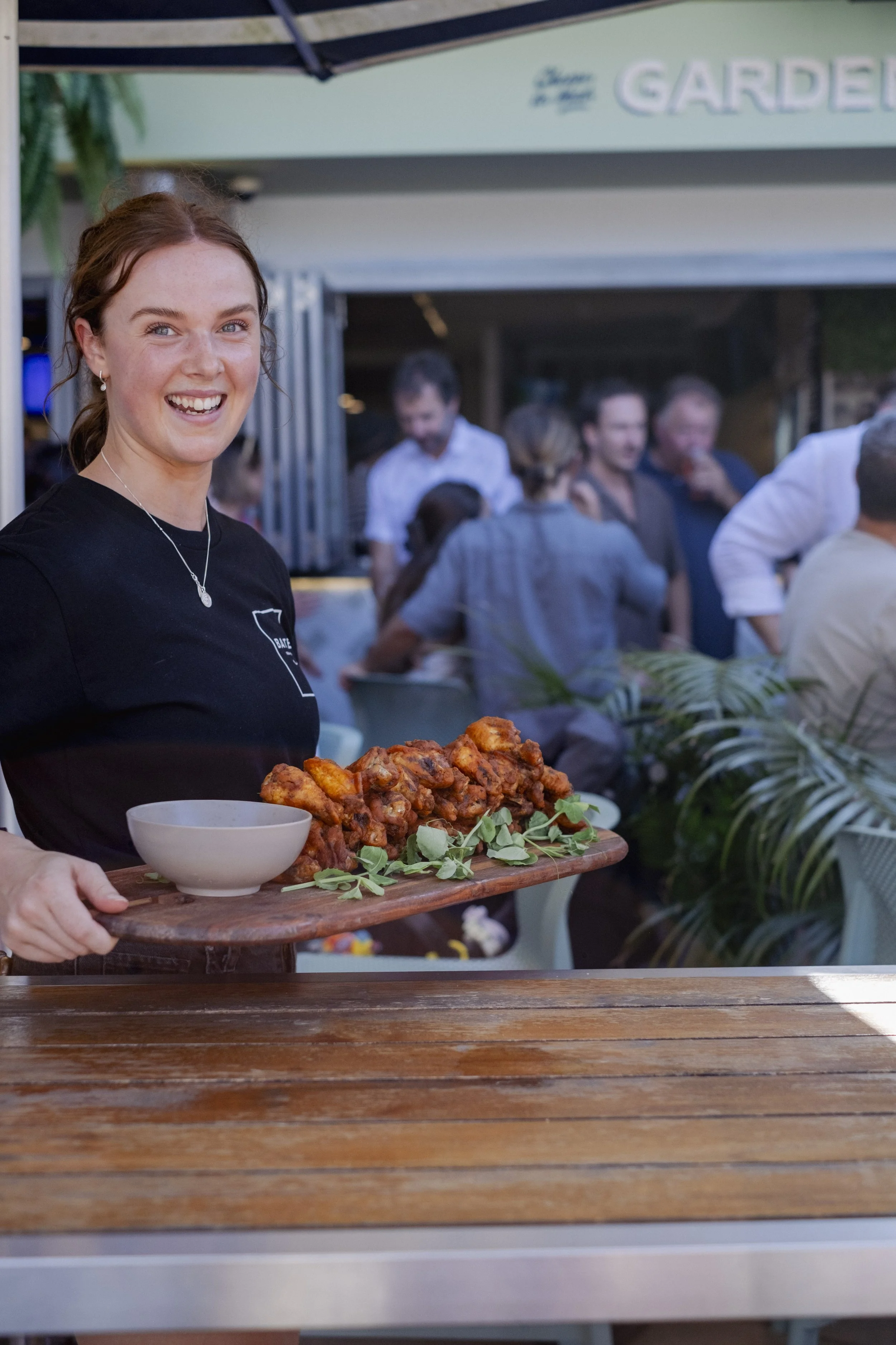 A smiling woman holding a wooden tray of fried chicken with lettuce garnishes, at an outdoor gathering or event.