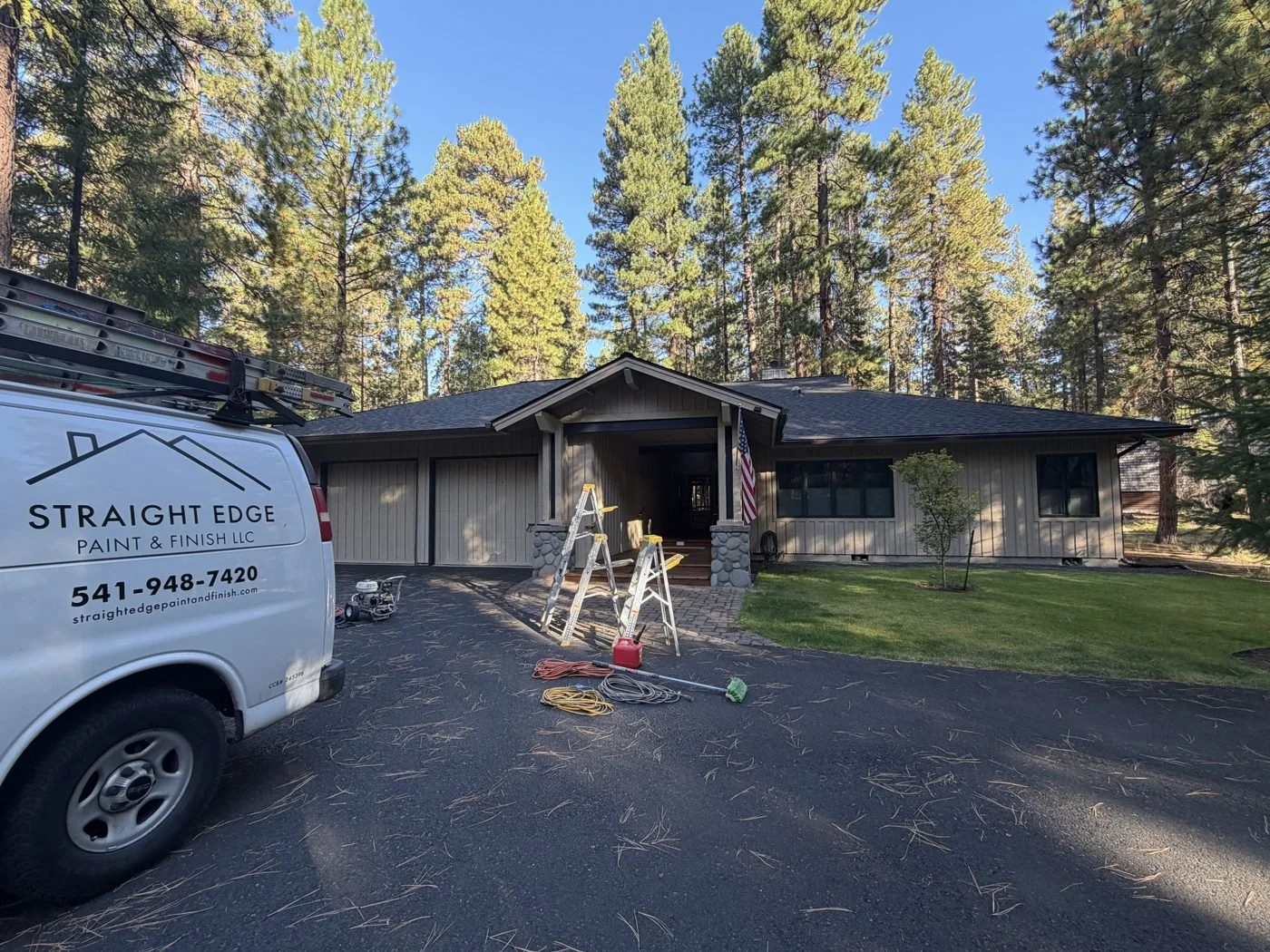 Single-story Black Butte Ranch home with light tan board-and-batten siding, two-car garage, and stone-column covered entry, surrounded by tall pine trees, before exterior painting