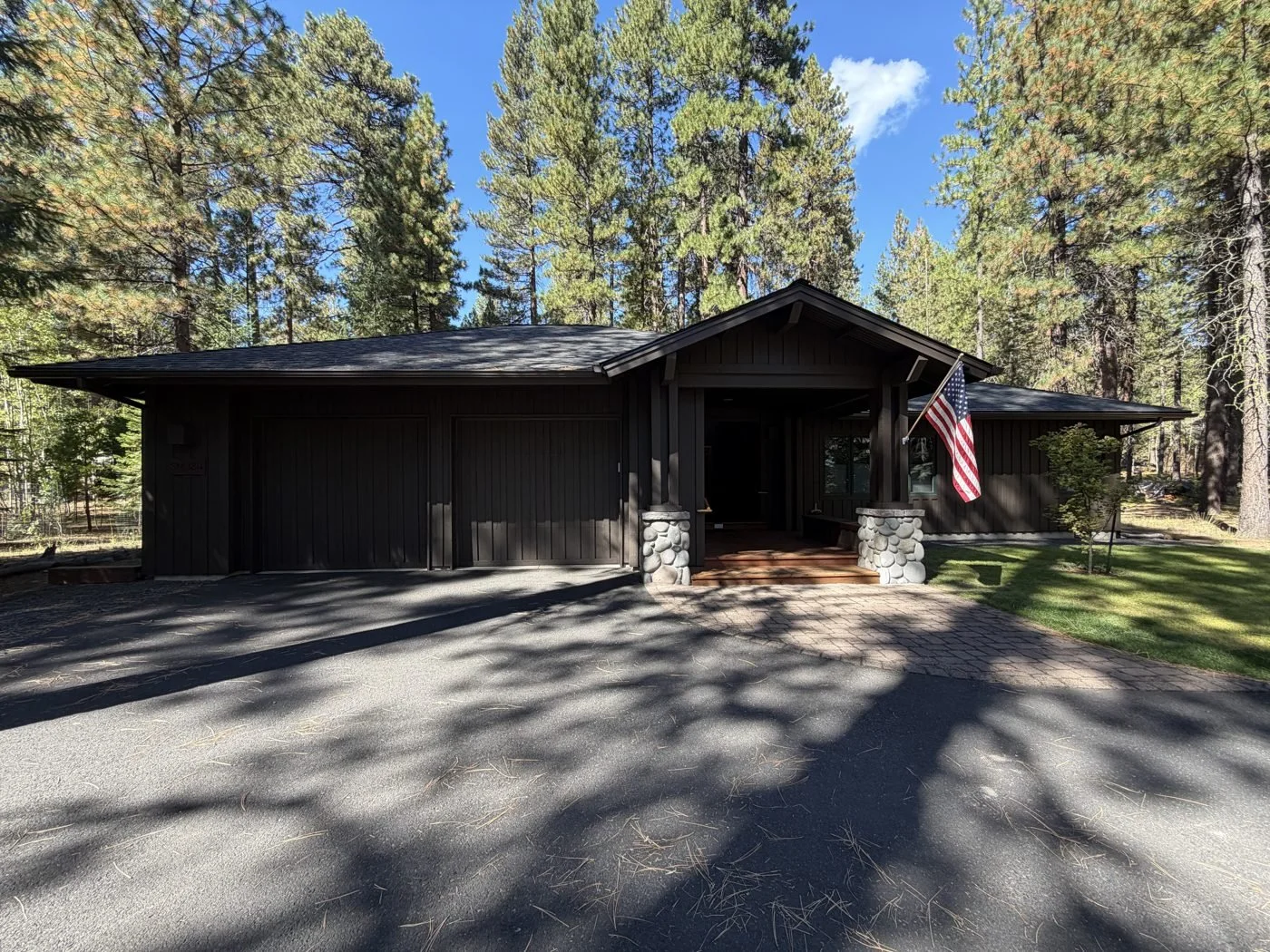 Same Black Butte Ranch home painted a dark brown, with board-and-batten siding, two-car garage, and stone-column covered entry among tall pine trees