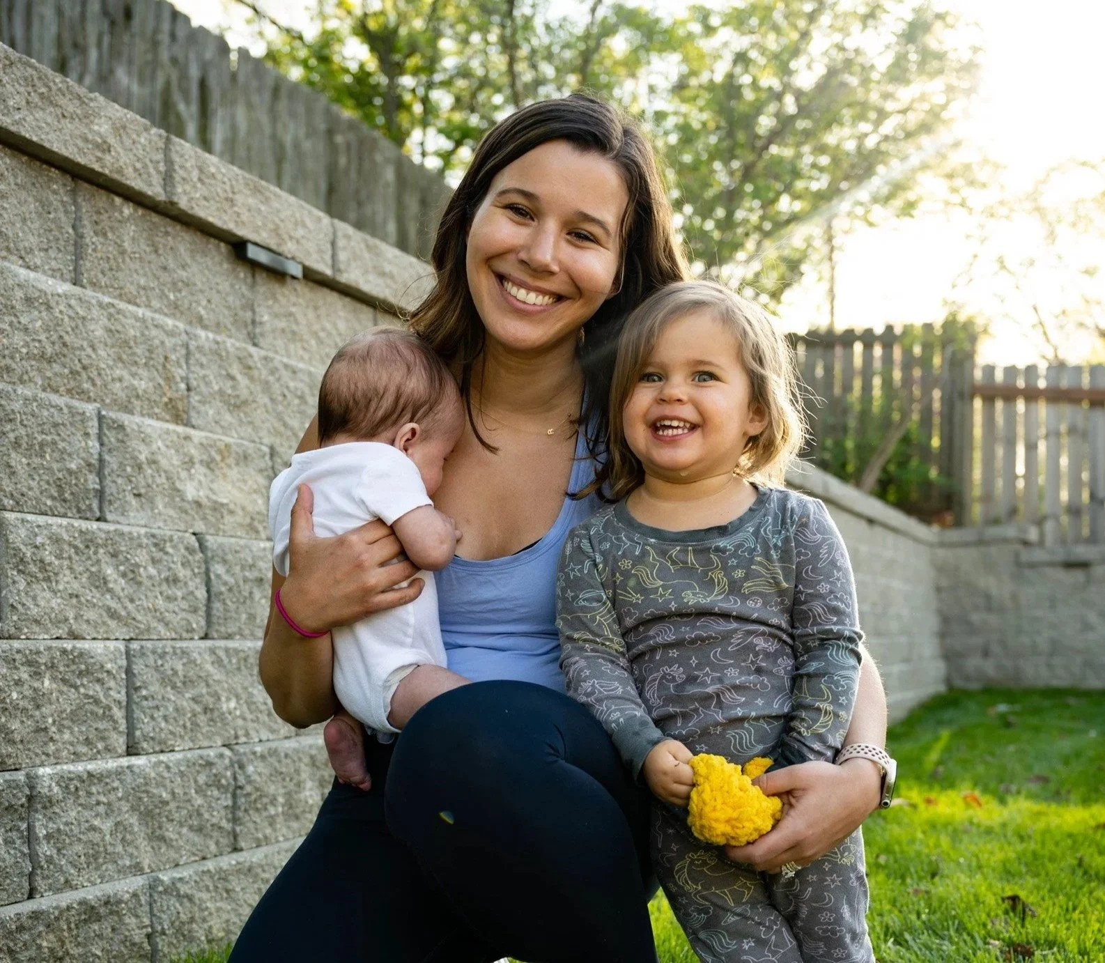 A smiling woman sitting outdoors with two young children, one holding a yellow toy, in a backyard with a stone wall and trees in the background.