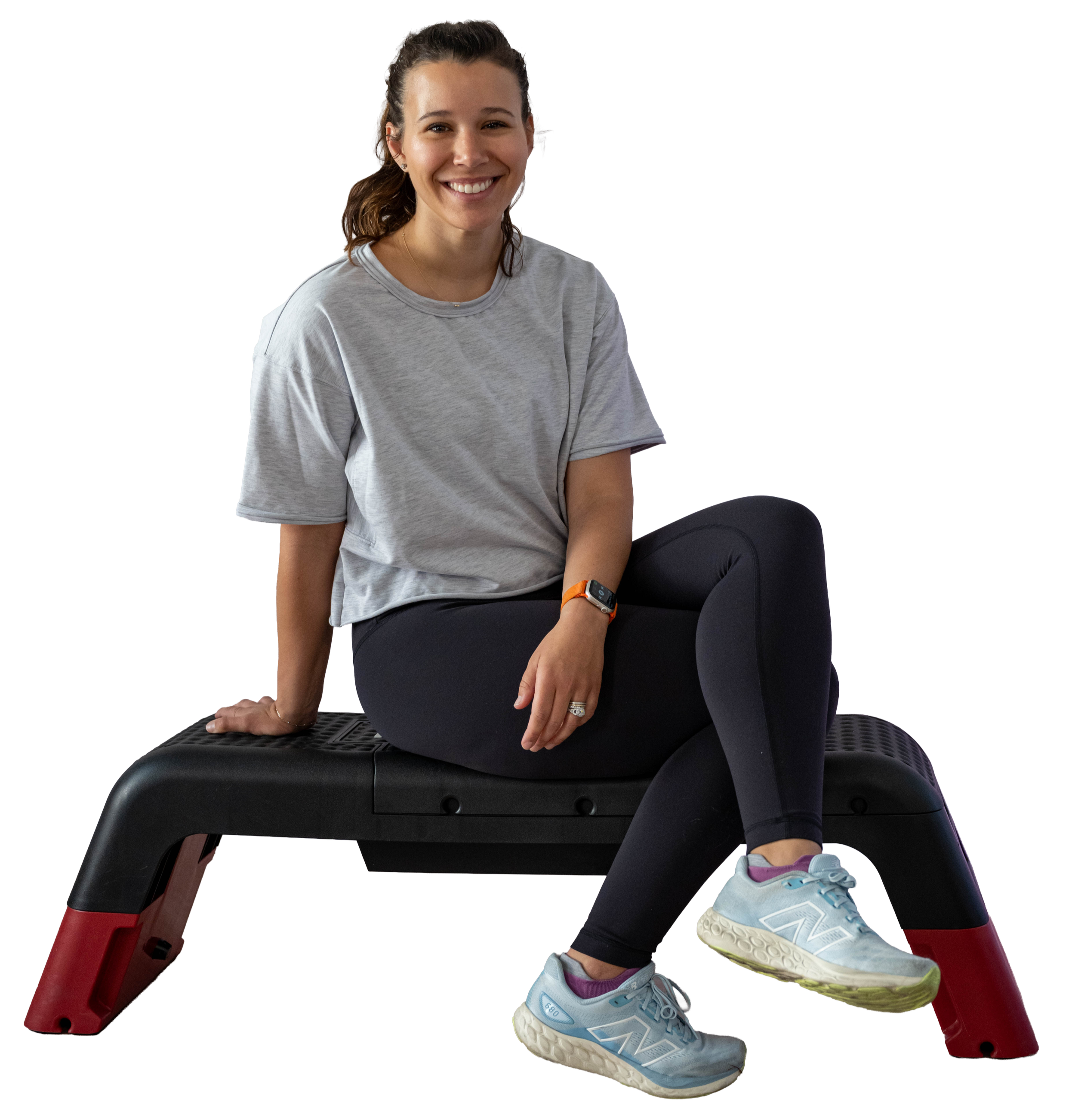 Natalie Exline in gray t-shirt and black leggings sitting on a black exercise bench, smiling, with athletic shoes, a fitness watch, and a ring.