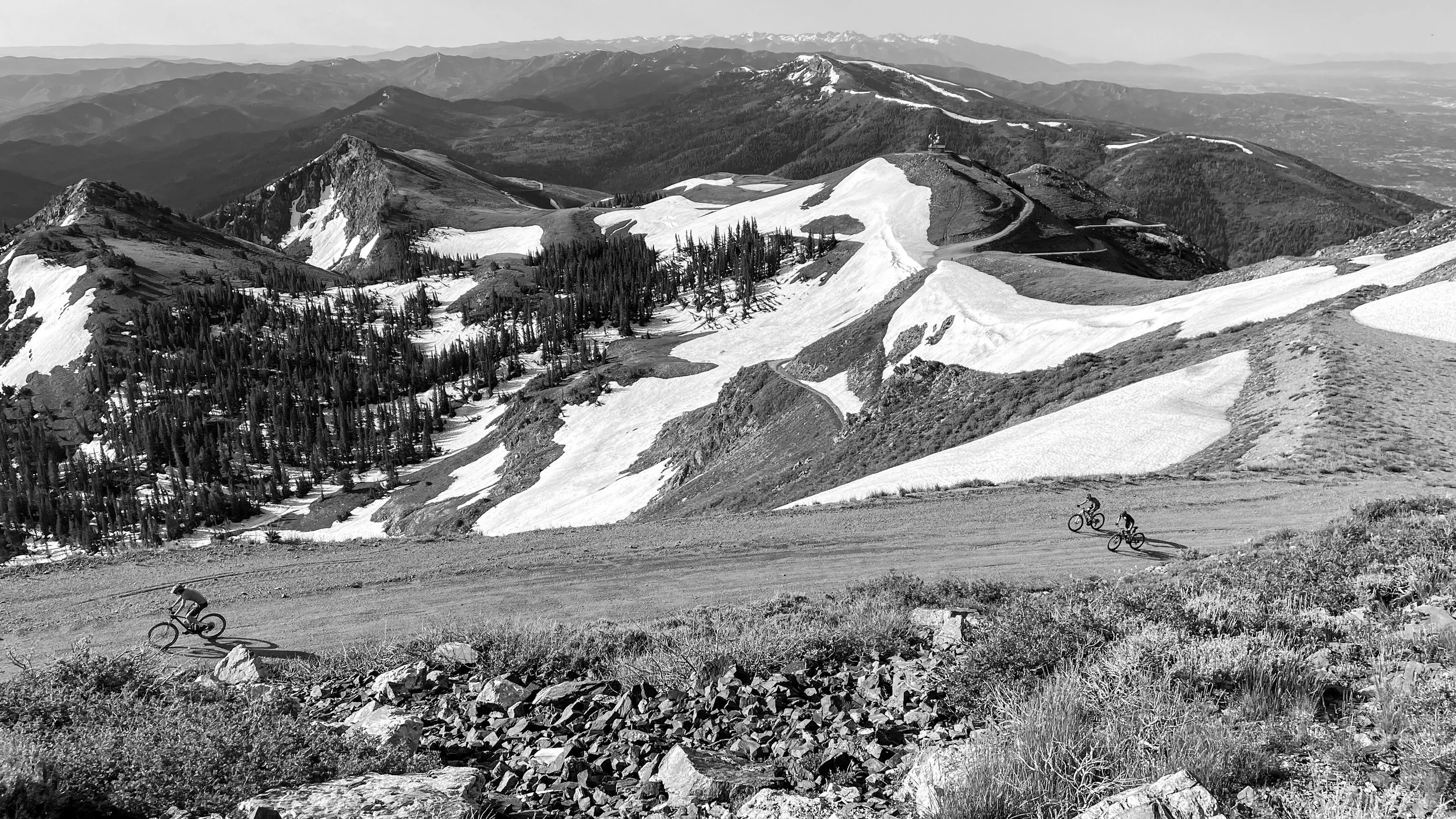 Mountain landscape in black and white with snow patches on peaks and slopes, several cyclists riding along a dirt road, dense forest on hillsides, distant mountain ranges.
