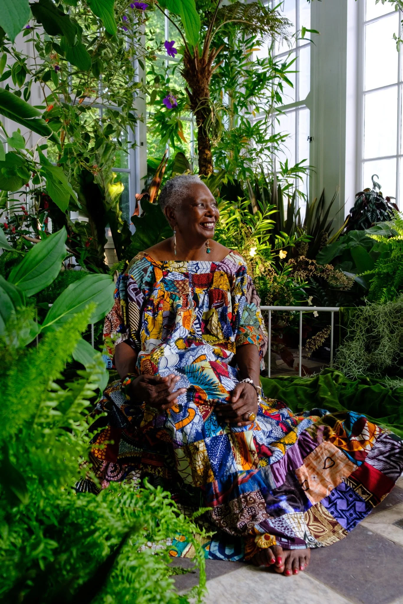 An elderly woman with gray hair, wearing colorful patterned dress and earrings, sitting among many green plants and flowers inside a bright conservatory with large windows, smiling and looking to her right.