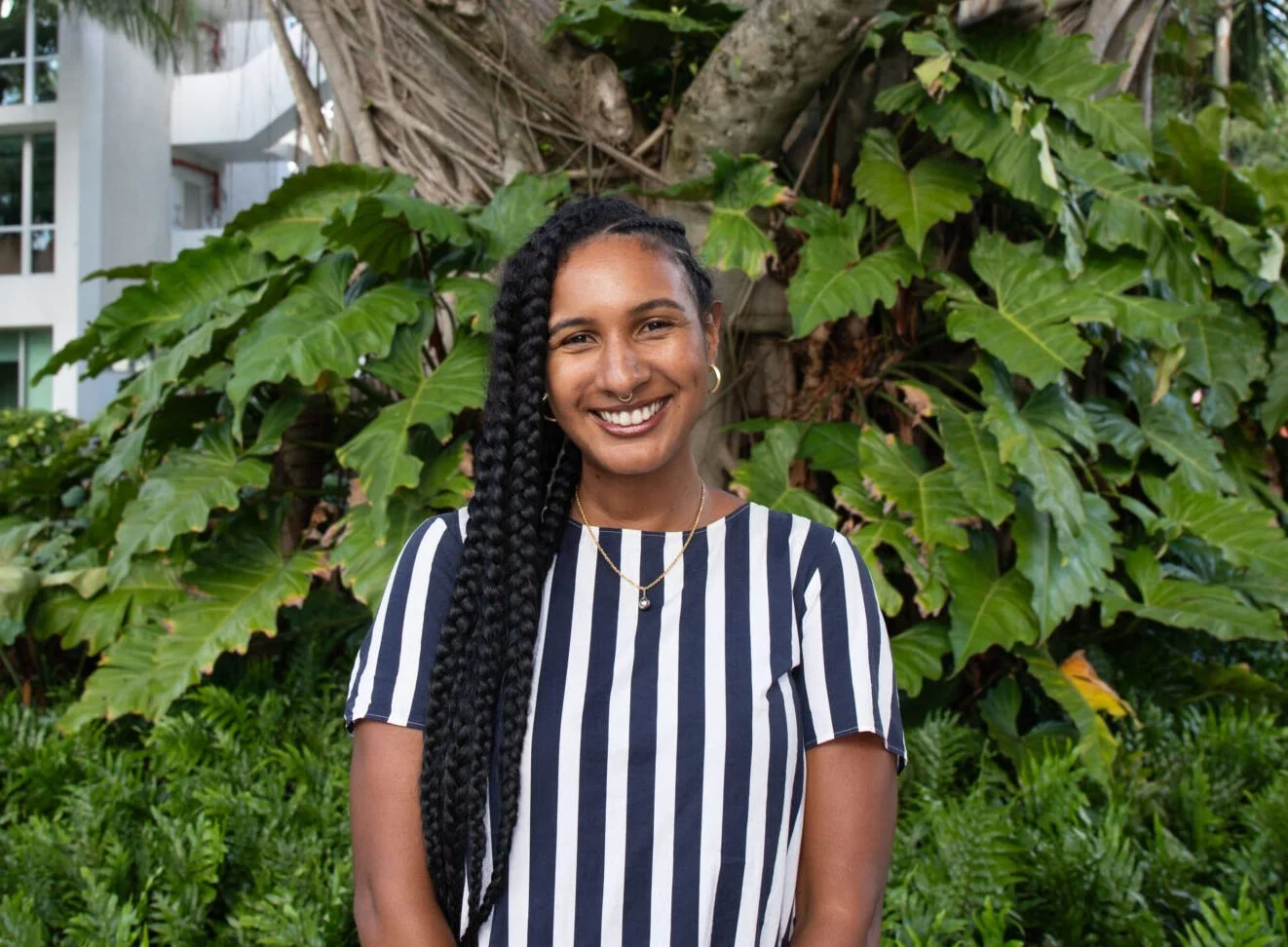 Marina Magloire, a Black woman with long braided hair smiling outdoors, wearing a black and white striped shirt and gold jewelry, standing in front of a lush greenery background.