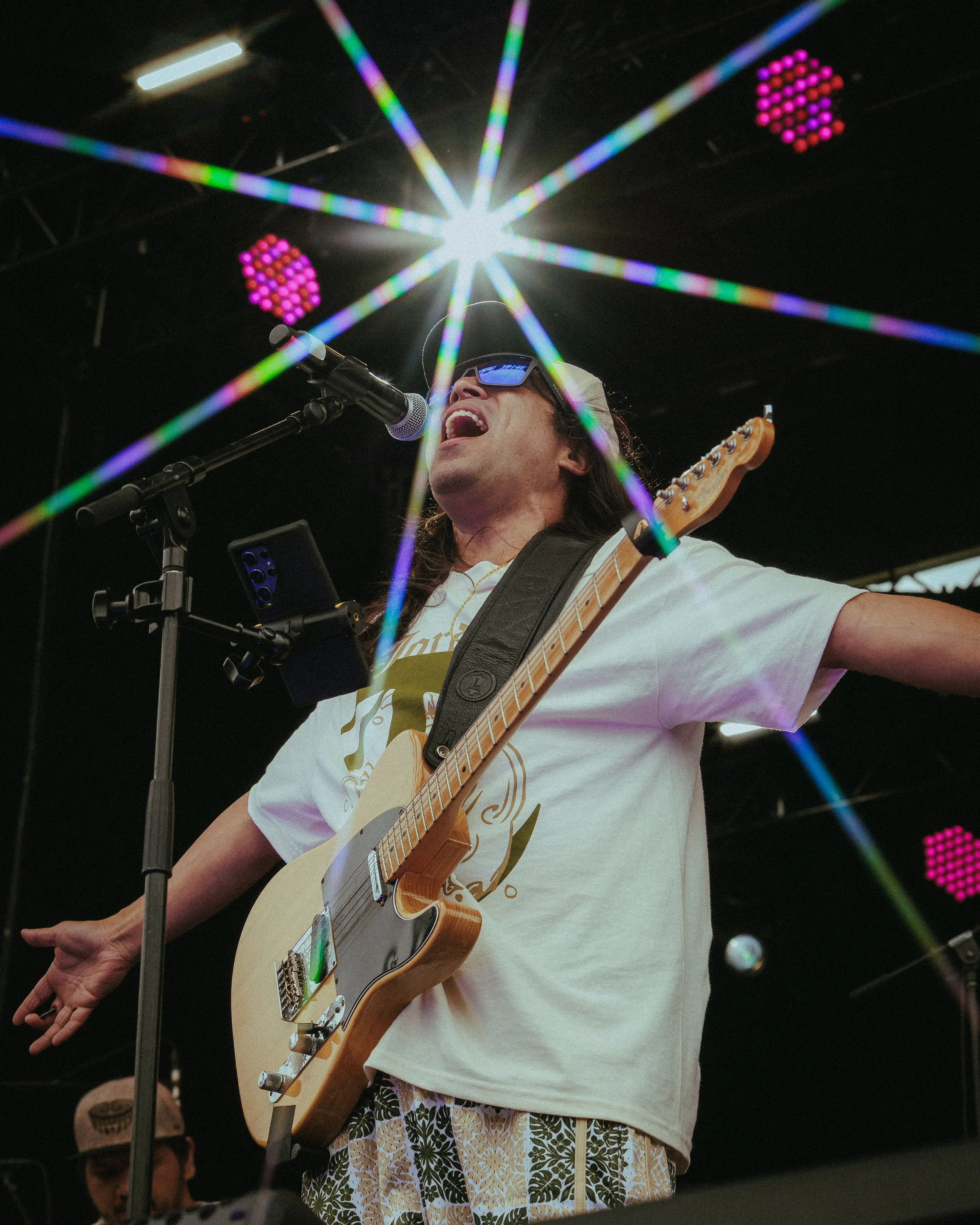 Musician singing into a microphone while playing electric guitar under colorful stage lights.