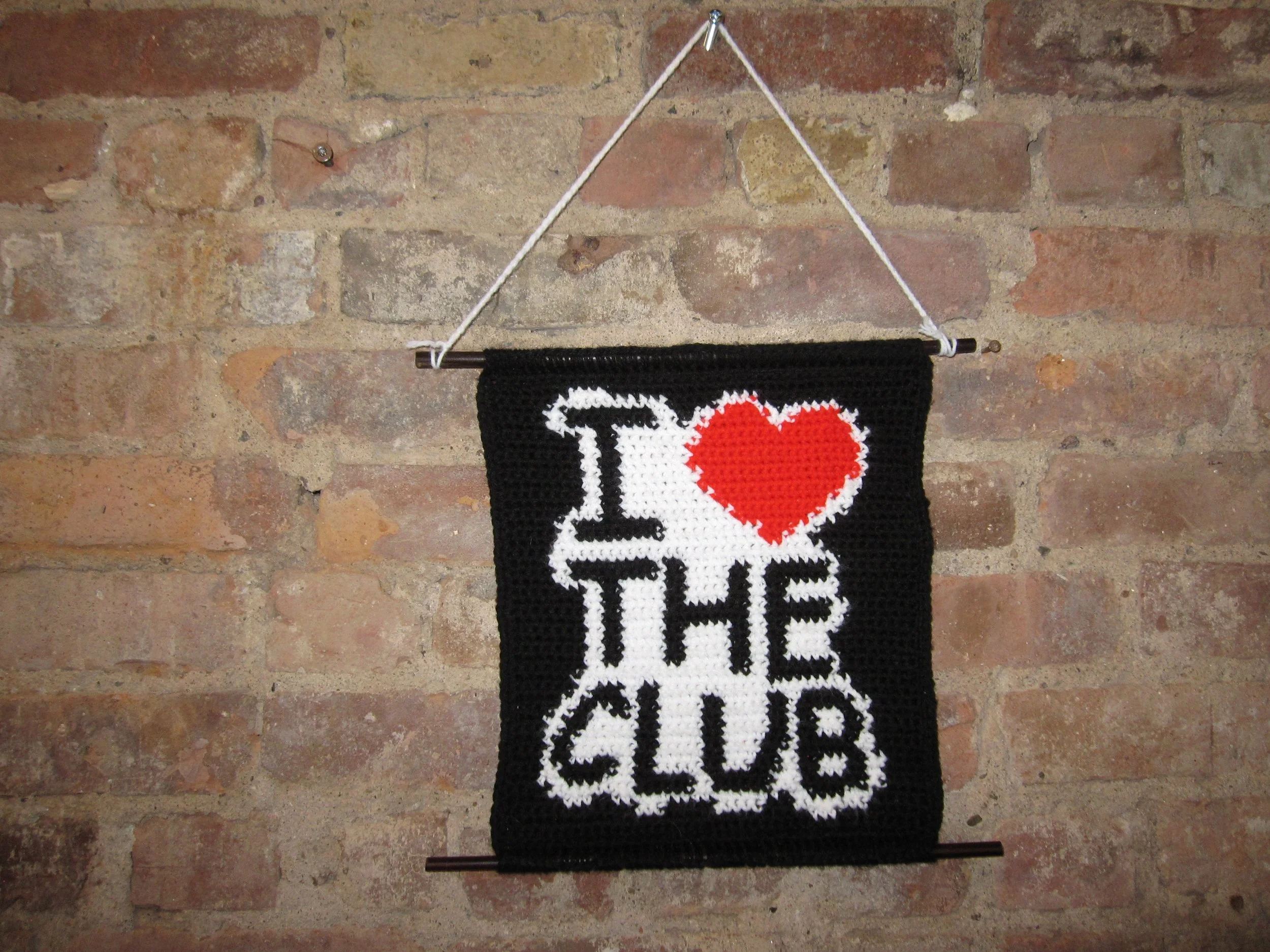 Crochet wall hanging with the phrase "I ❤ THE CLUB" against a brick wall background.
