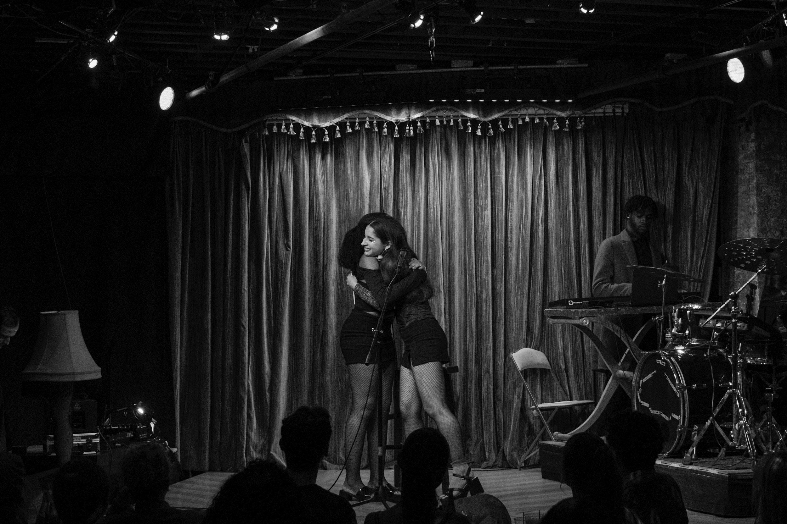 Two women hugging on a stage with musical instruments in the background, including a keyboard, drum set, and a microphone, in front of a curtain.