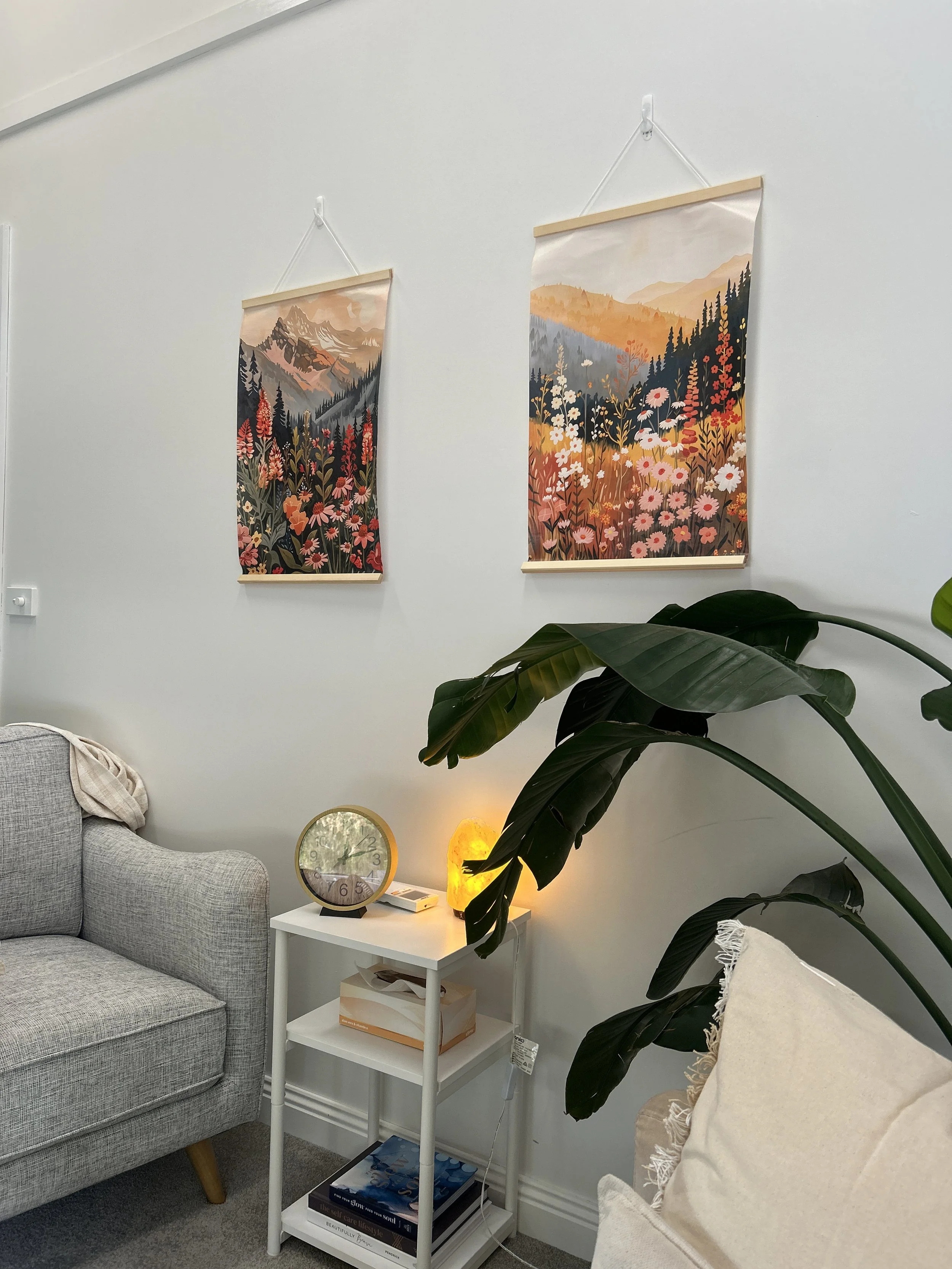Counselling room with two colorful landscape hanging art pieces on the white wall, a gray fabric sofa, a white side table with a round clock, a salt lamp, a box of tissues, a potted plant, and a cream cushion.
