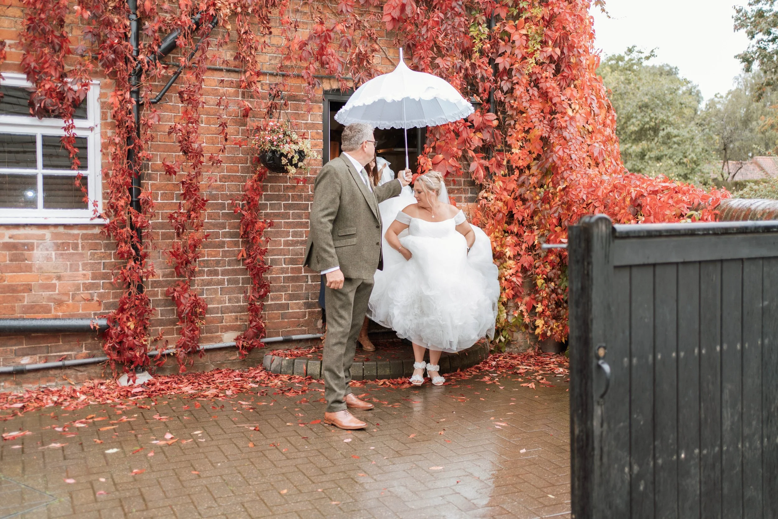 A bride in a white wedding dress looks down while standing outside a brick building with red ivy. A man holding a white umbrella over her is standing nearby. The ground is wet with fallen leaves, and there is a black trash bin in the foreground.