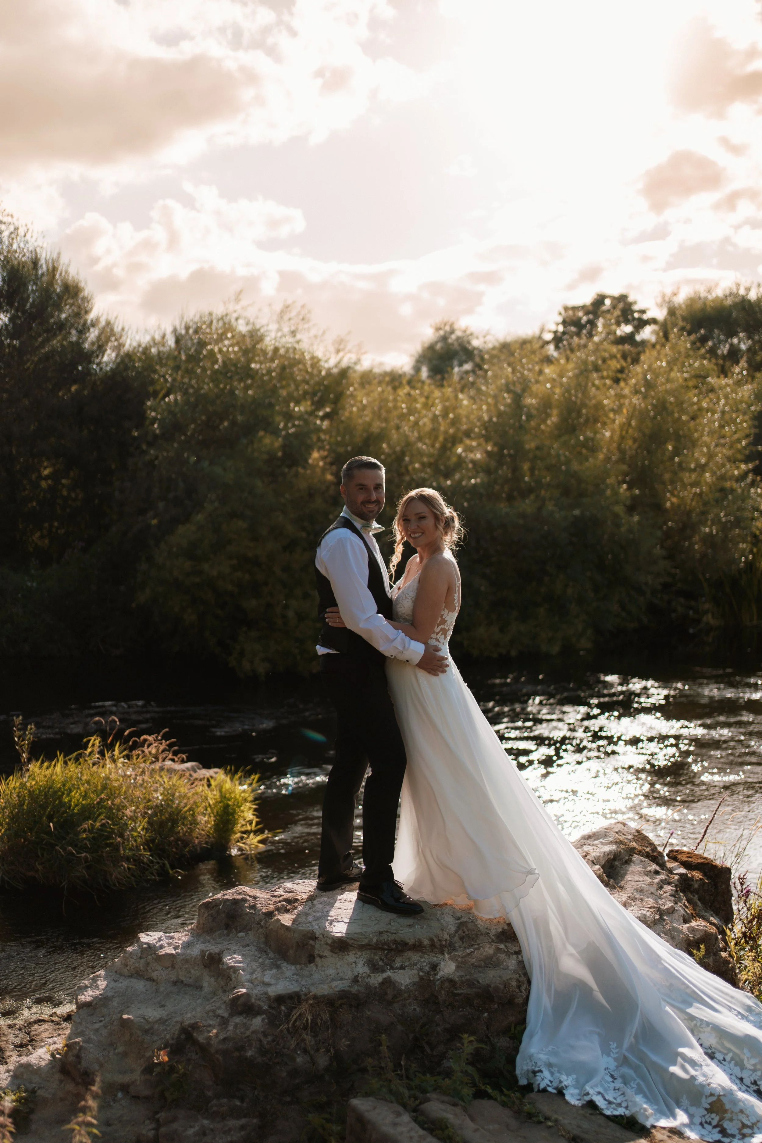 A newlywed couple stands on a large rock near a creek, with a backdrop of trees and a cloudy sky, during sunset.
