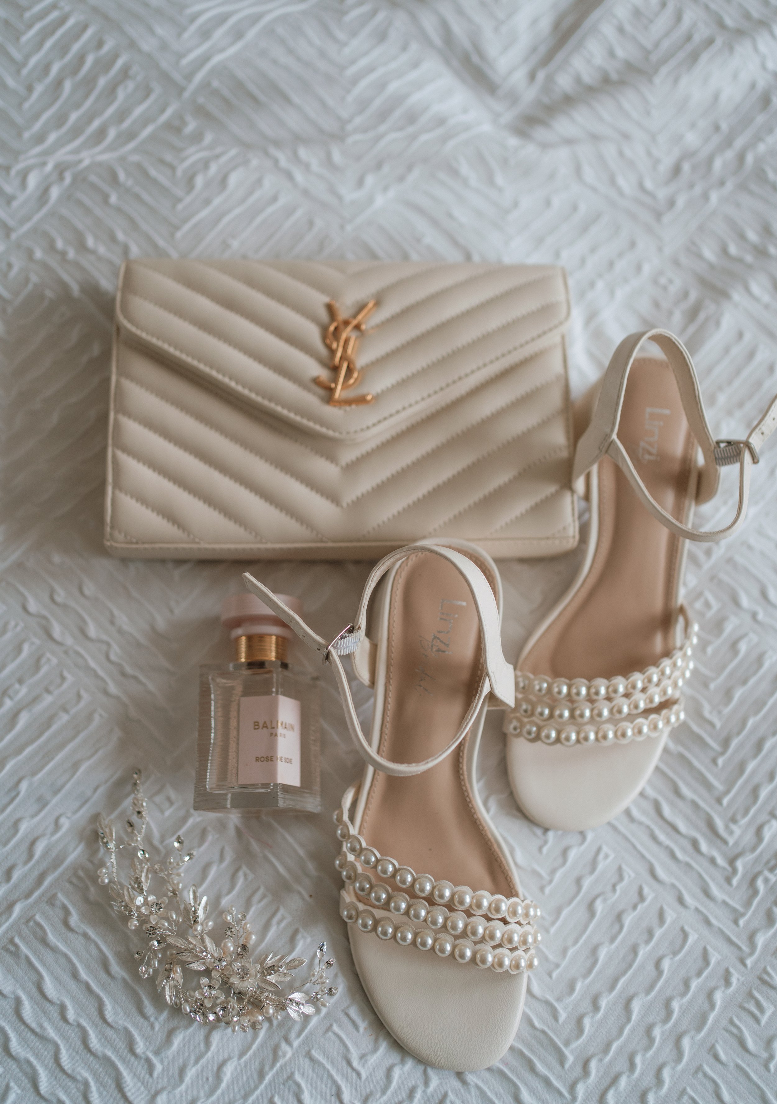 An arrangement of white and cream-colored accessories, including a quilted wallet with a gold YSL logo, a pair of white high-heeled shoes with pearl embellishments, a pearl bracelet, a pearl necklace, and a bottle of Balmain Paris perfume on a textur