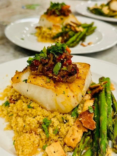 Seared white fish fillet with garnished tomato relish, served with couscous and grilled asparagus on a white plate.