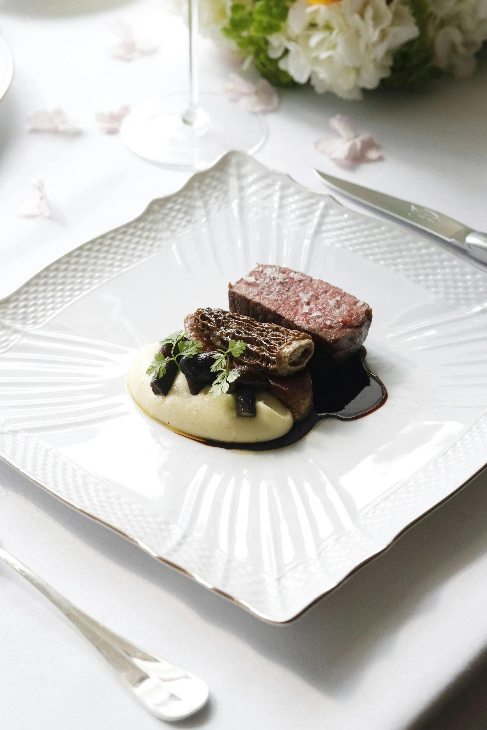 A plated gourmet dish features a round beef or steak fillet garnished with fresh microgreens, served on a bed of bright yellow mashed potatoes, with a drizzle of dark berry or wine sauce around the base.