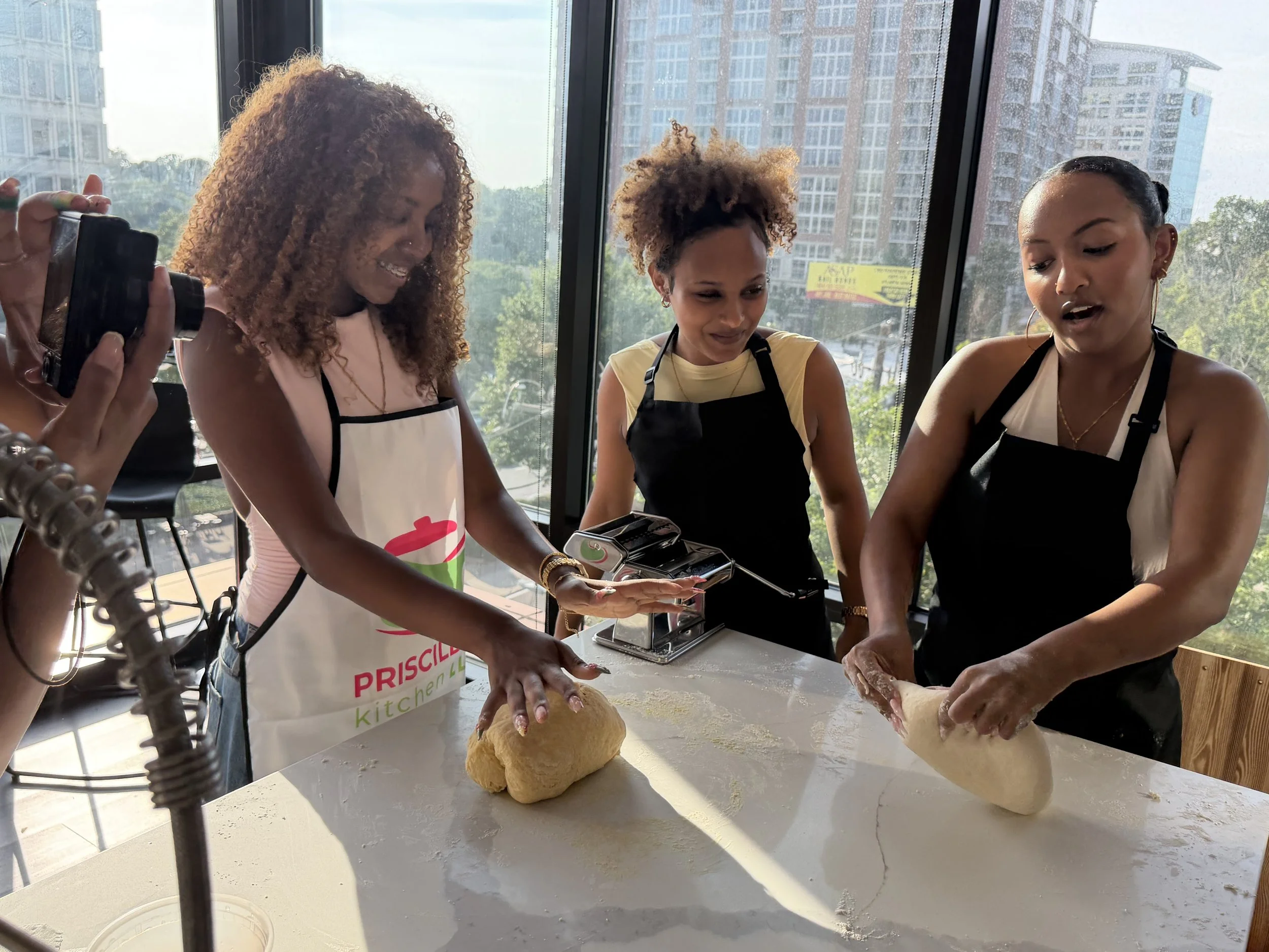 Group of people wearing aprons with 'Priscilla's Kitchen' logo, preparing food together in a kitchen, smiling, with some holding knives and others chopping or serving food.