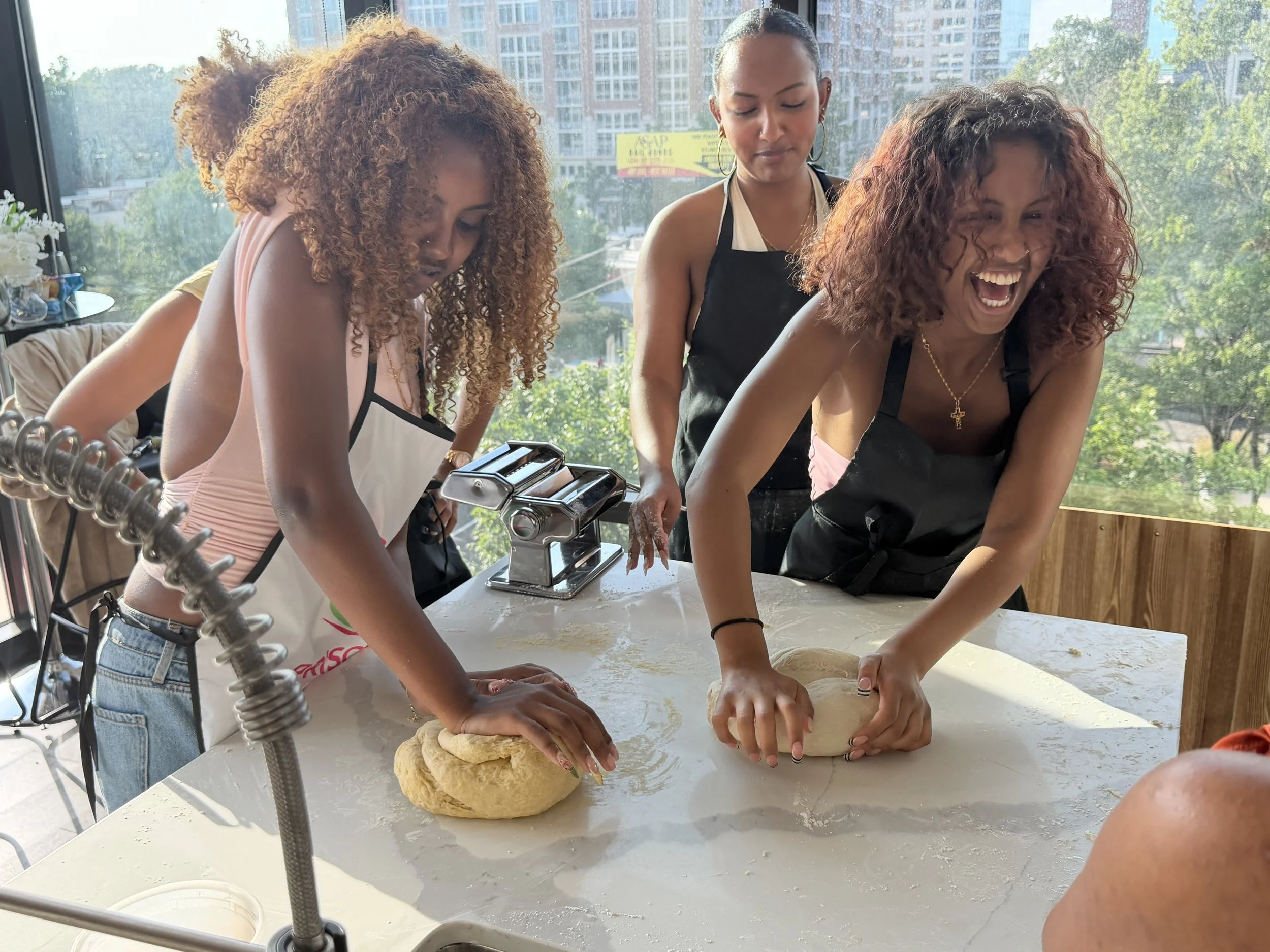 Group of people wearing aprons with 'Priscilla's Kitchen' logo, preparing food together in a kitchen, smiling, with some holding knives and others chopping or serving food.