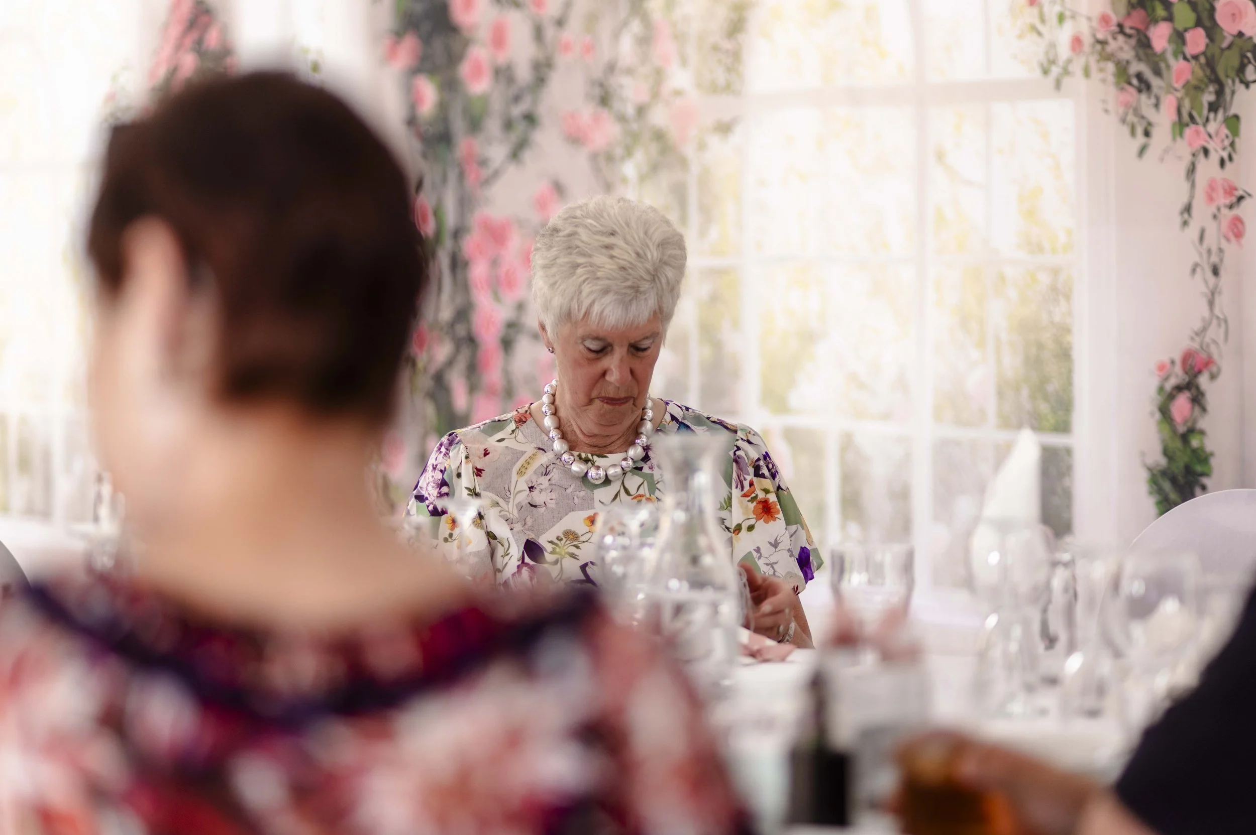 An elderly woman with short gray hair looking down, seated at a table with a floral tablecloth, surrounded by glassware and a floral wallpaper background at a wedding.