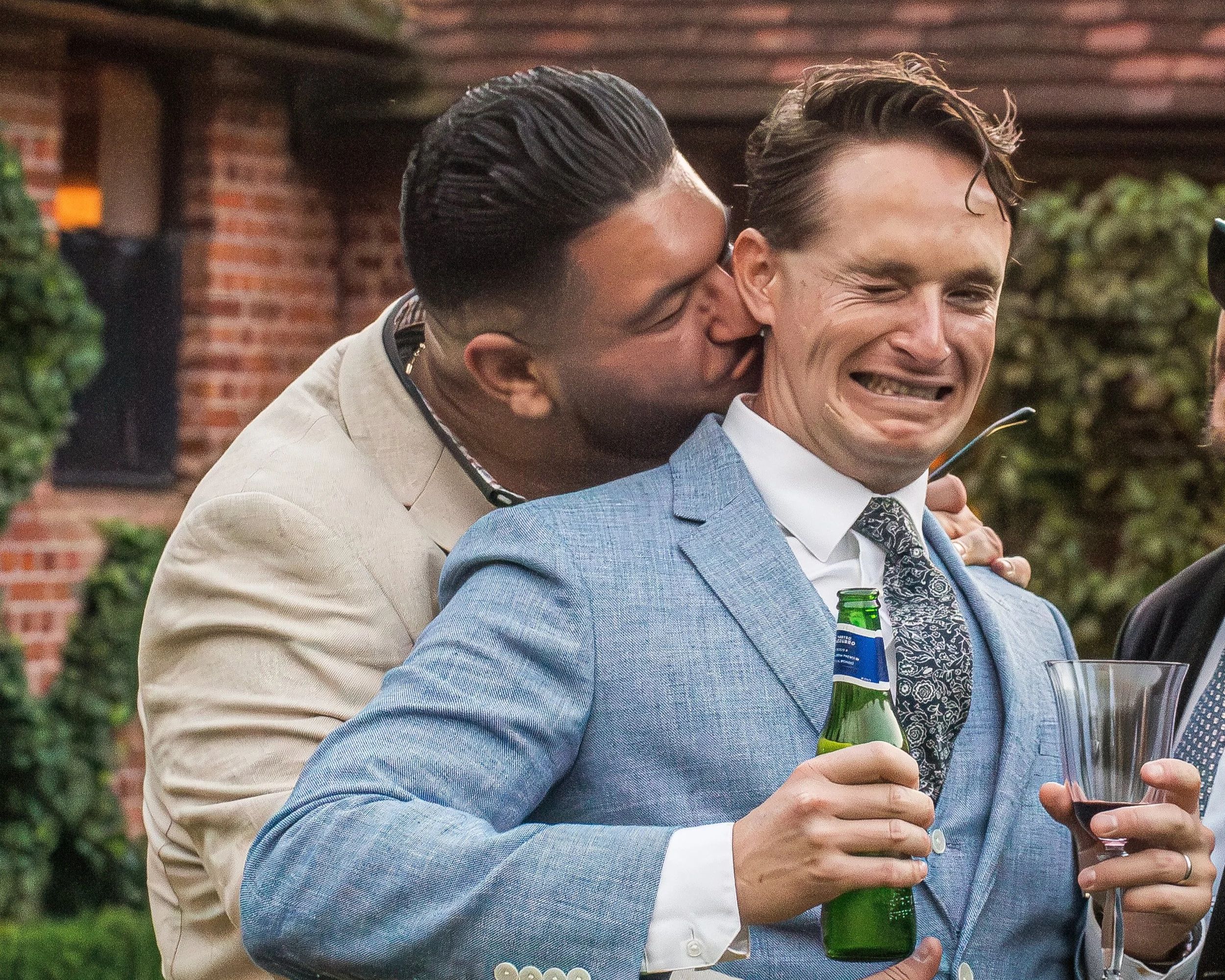 Two men at a wedding, one in a light blue suit with a drink, and the other in a beige suit kissing him on the cheek.