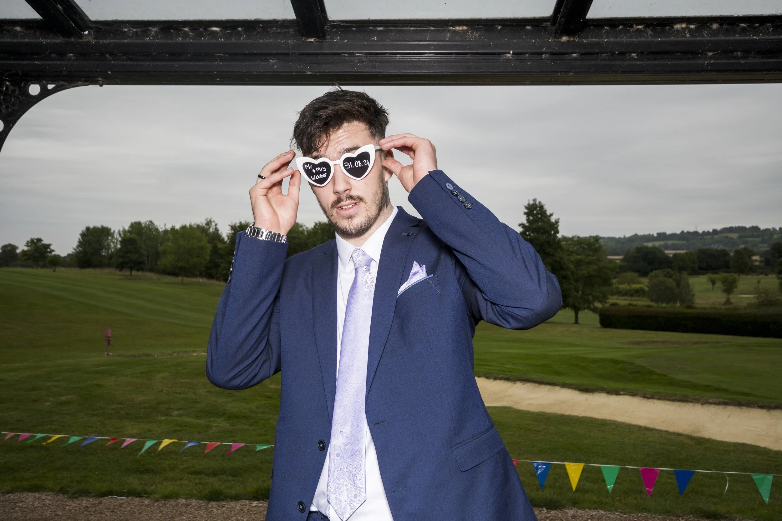 A man in a blue suit holding heart-shaped sunglasses with details for a wedding, standing outdoors with a golf course and trees in the background.
