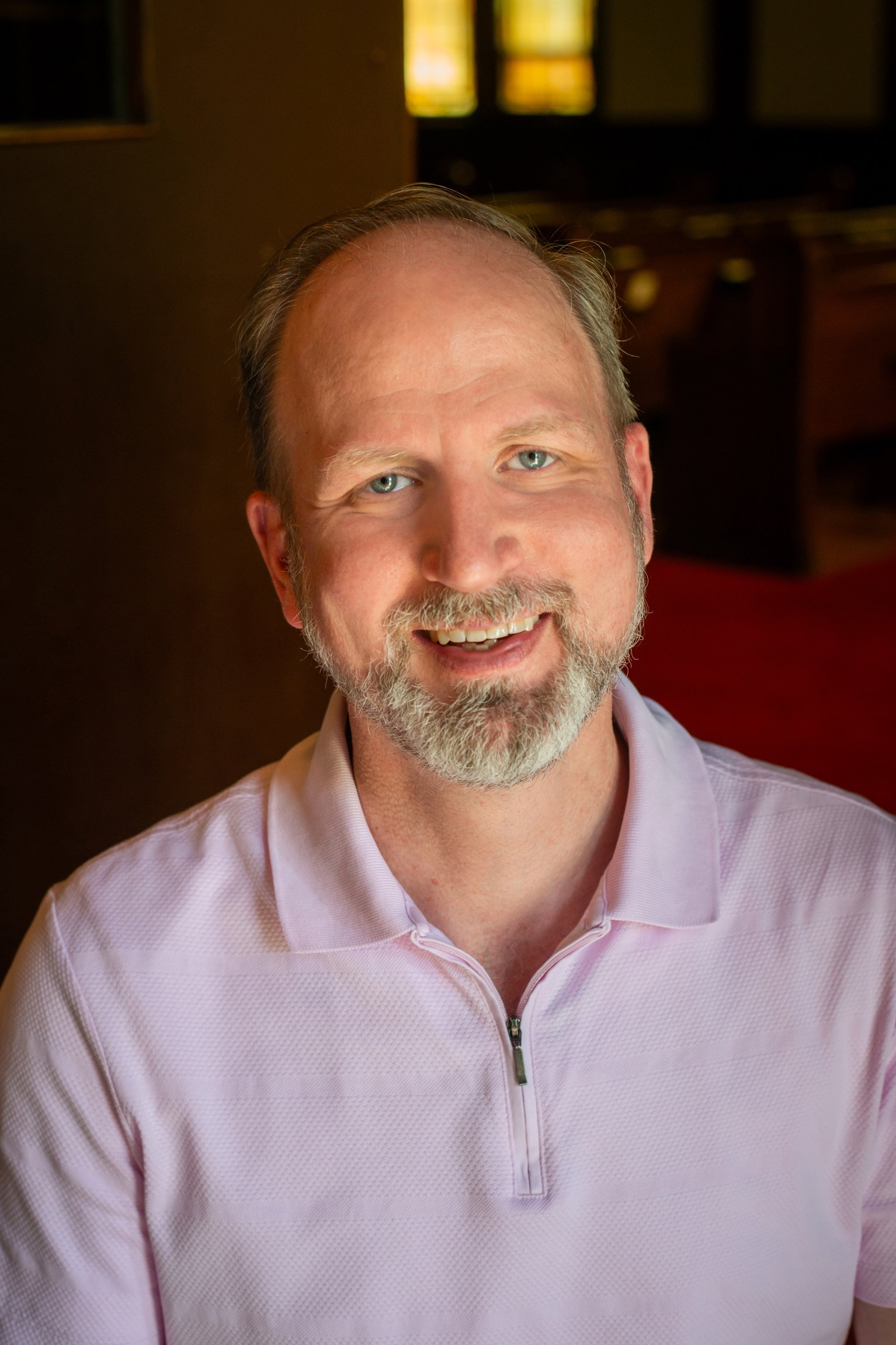 A smiling man with light skin, blue eyes, short brown hair, wearing a blue button-up shirt, standing against a dark grey background.