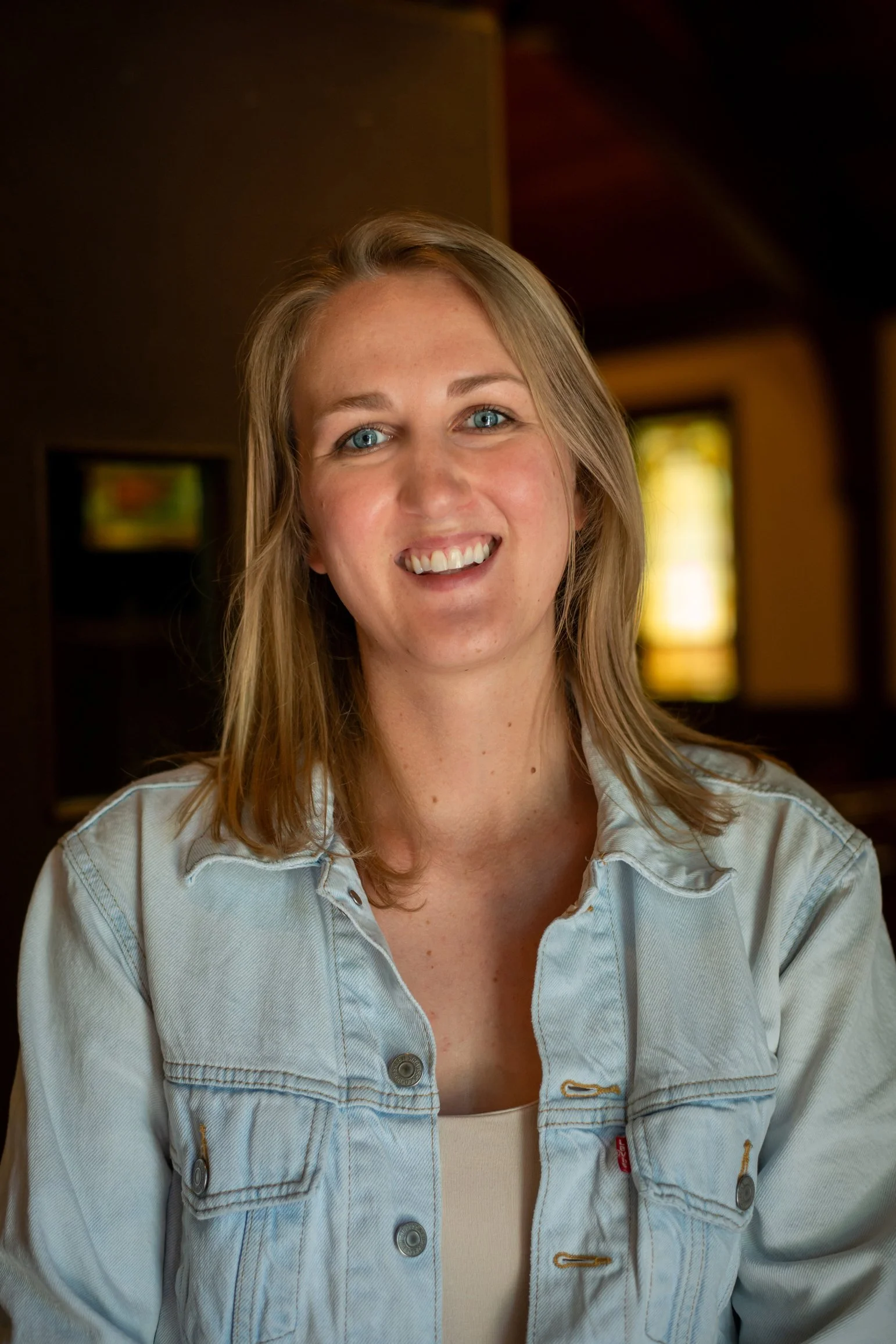 A smiling woman with wavy blonde hair wearing a black and white patterned top, standing against a plain white background.