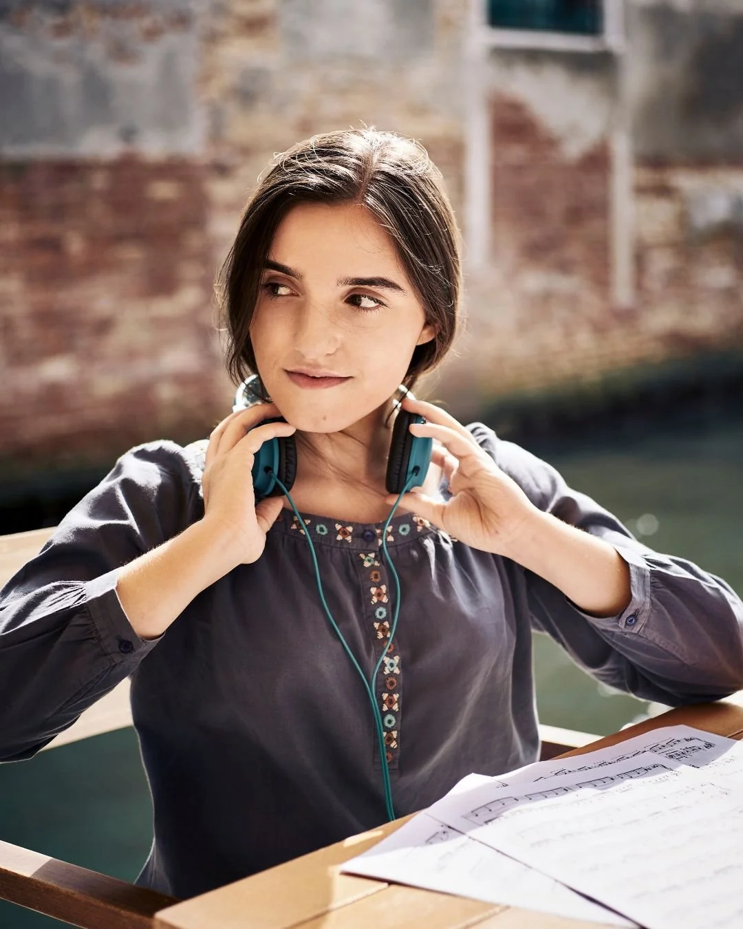 Una donna giovane con i capelli castani scuri si sta preparando a suonare uno strumento musicale con le cuffie, seduta a un tavolo con delle partiture musicali davanti a lei, in un ambiente all'aperto vicino a un canale o un fiume.