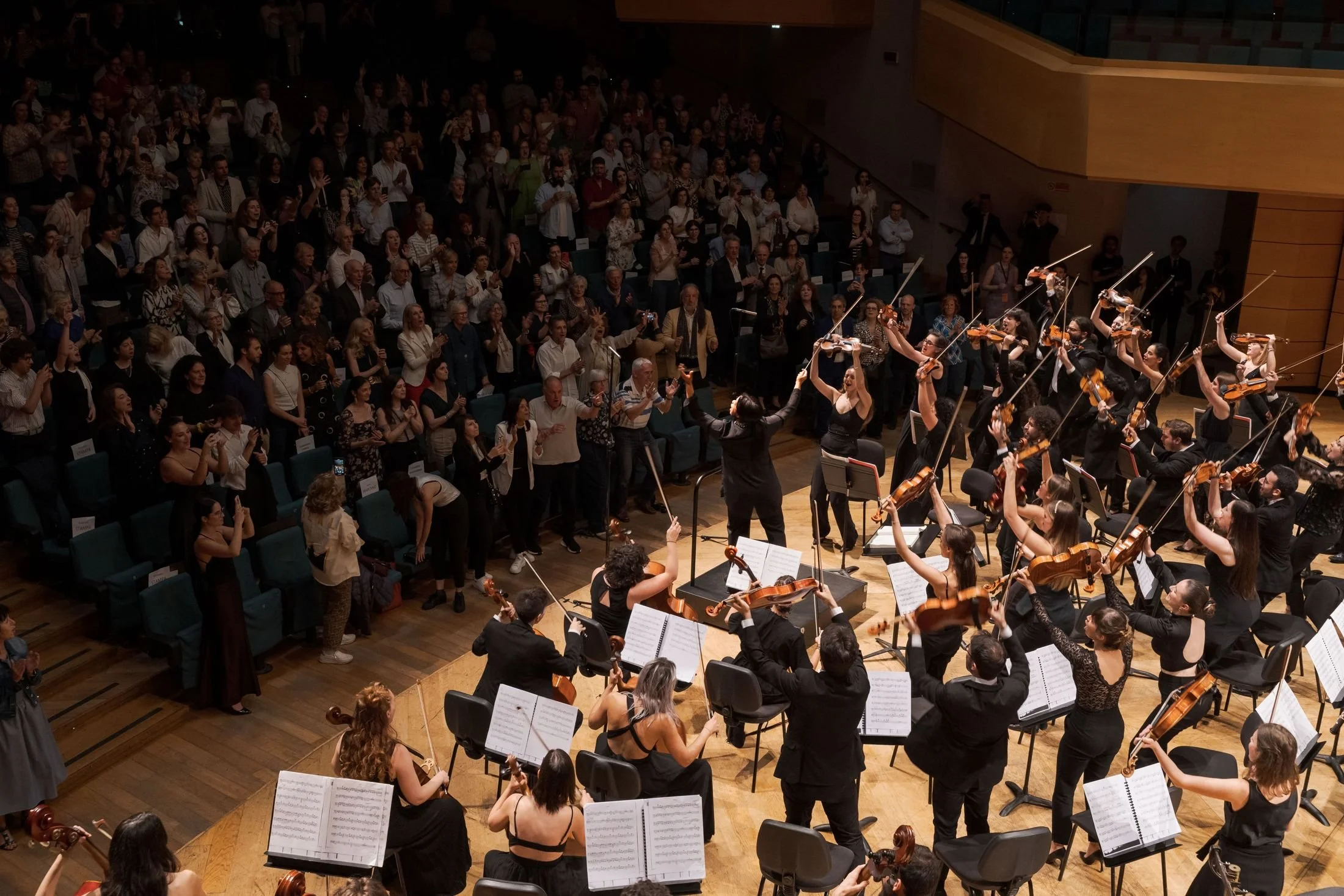 Un'orchestra di musicisti che suonano nel pubblico di un teatro o sala da concerto, con un pubblico presente che applaude.