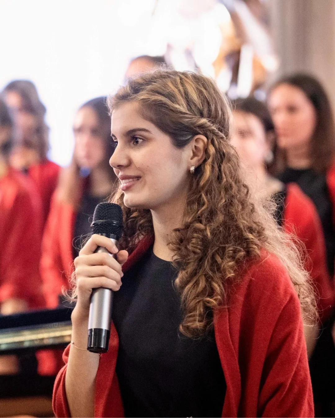 Una giovane donna con capelli ricci, vestita con una maglietta nera e una giacca rossa, tiene un microfono e sembra partecipare a un evento o a un'intervista, con altre persone sullo sfondo.
