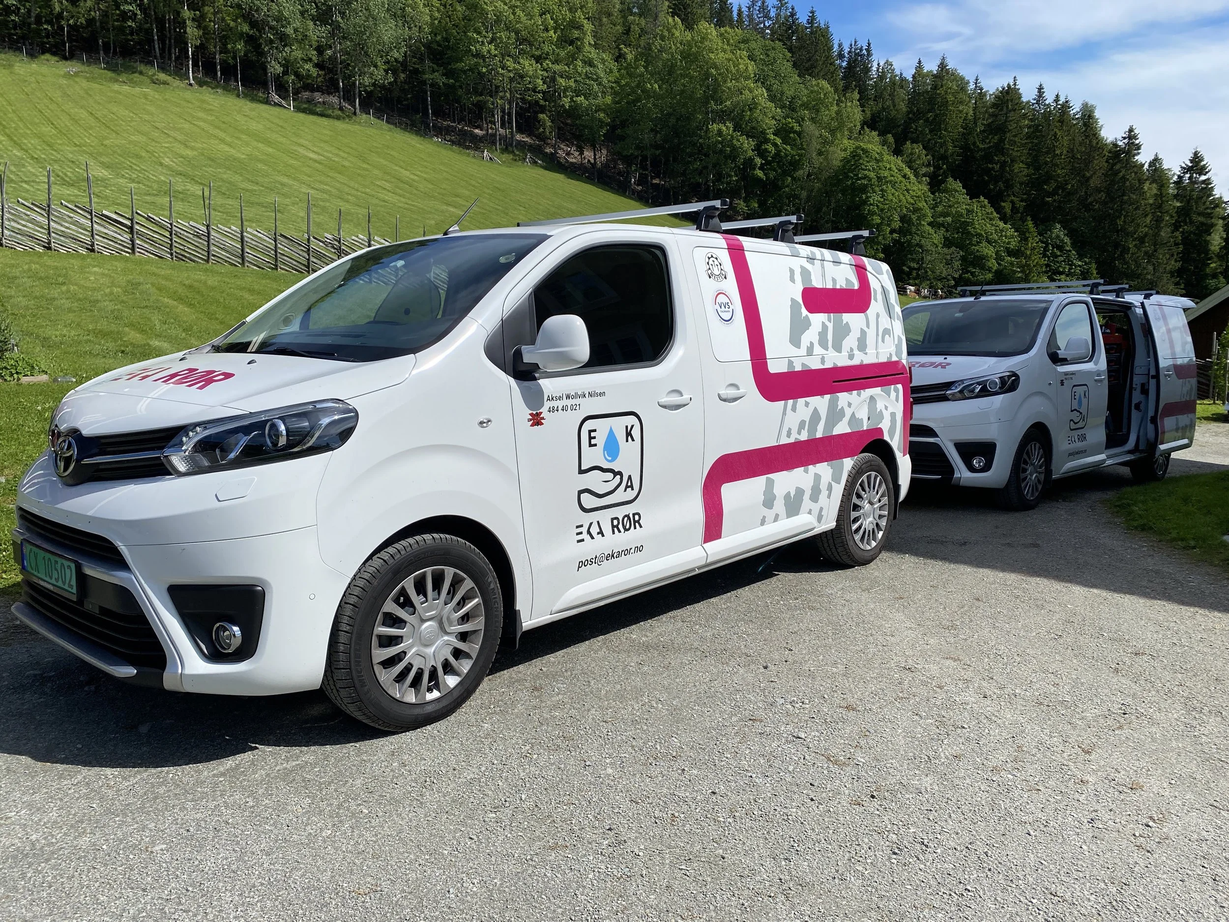 To hvite servicebiler med logoer og namn på, parkert på en grusvei, med grønne åser og trær i bakgrunnen.