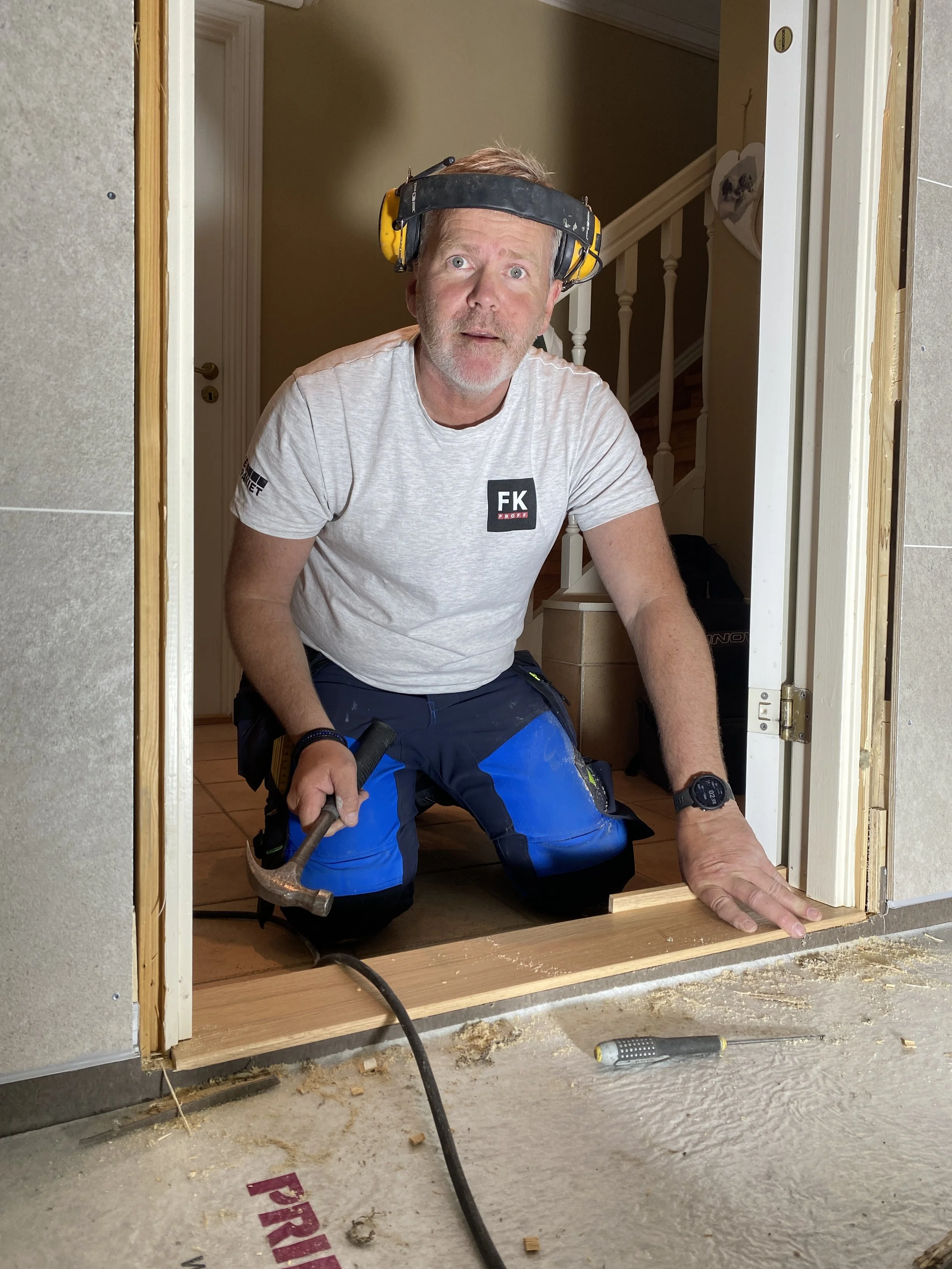 En mann i arbeid med å installere en dørkarm, iført t-skjorte, arbeidsbukse og verneutstyr, på et innendørs byggested.