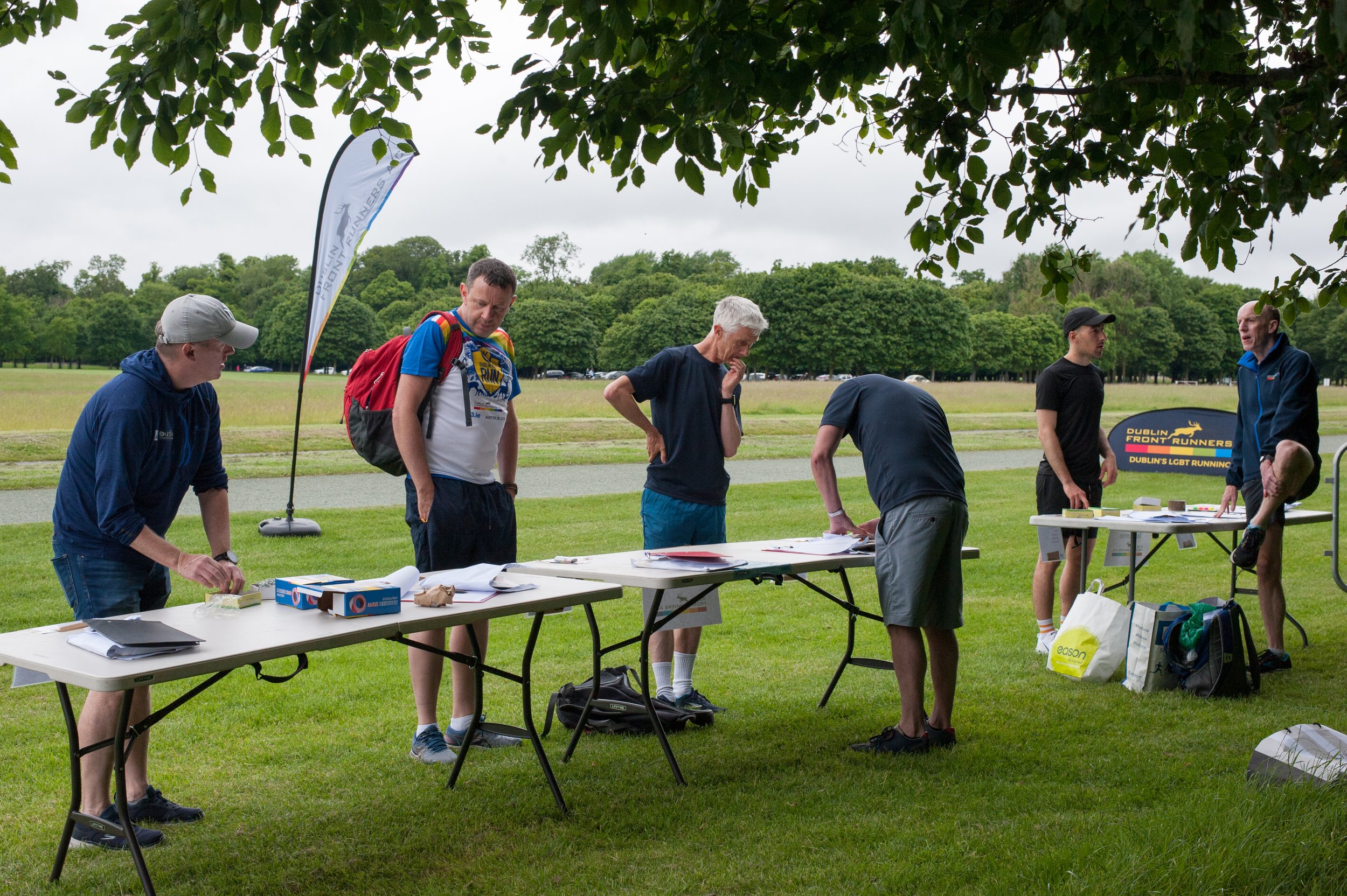 People standing at tables outdoors, possibly preparing for a running event. Signs mention "Dublin Front Runners," indicating a running group, with bags and documents on the tables. Trees and grass are visible in the background.