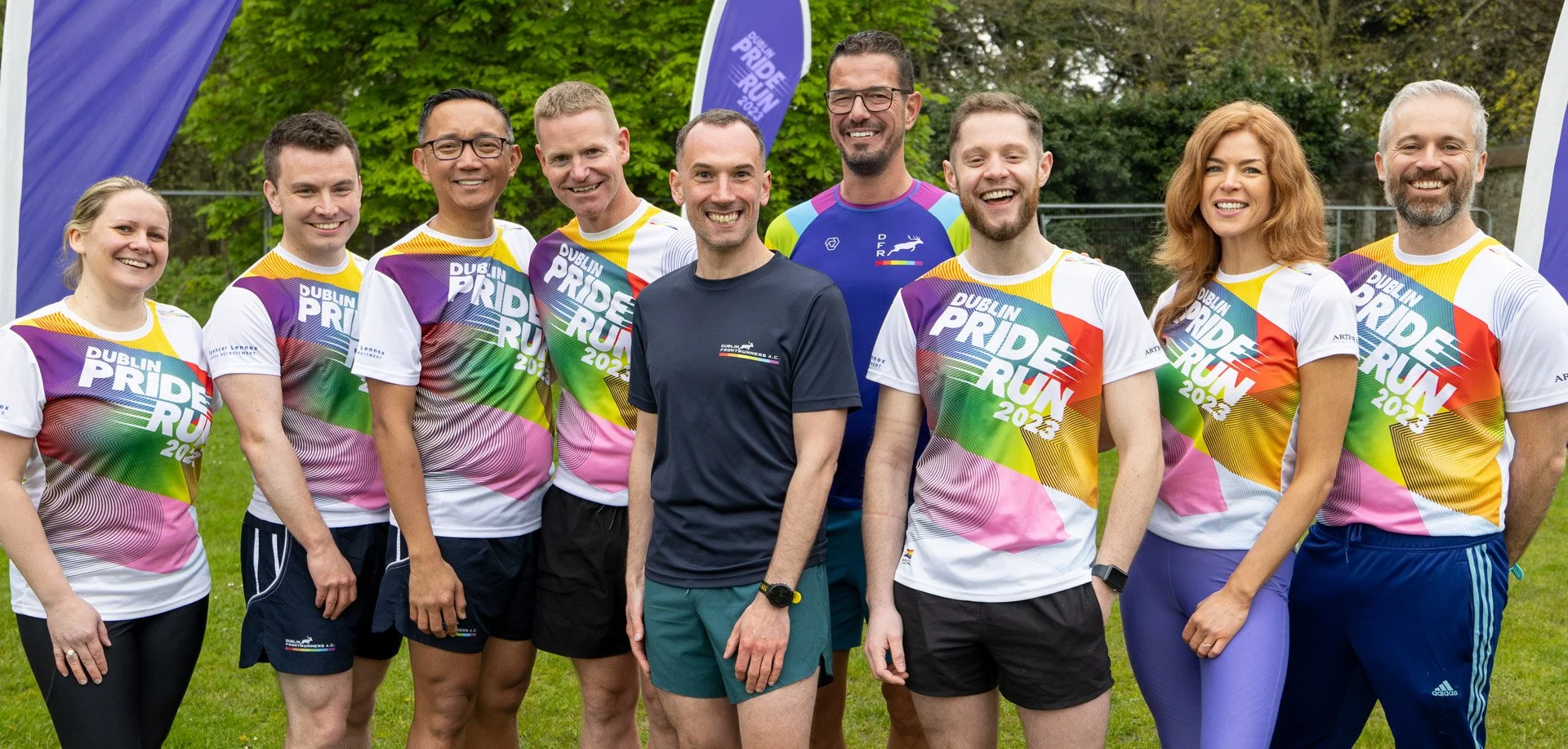 Group of people wearing colorful 'Dublin Pride Run 2023' T-shirts standing outdoors, smiling, with greenery in the background.