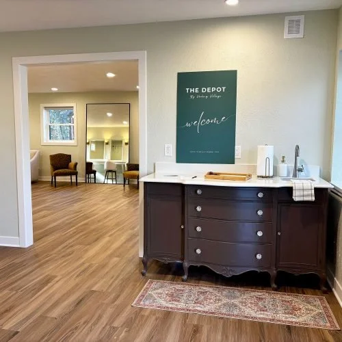 Interior of a room with a dark wood sideboard, welcome sign on the wall, and a small kitchen area with a sink and tea-making supplies. In the background, a seating area with chairs and a mirror.