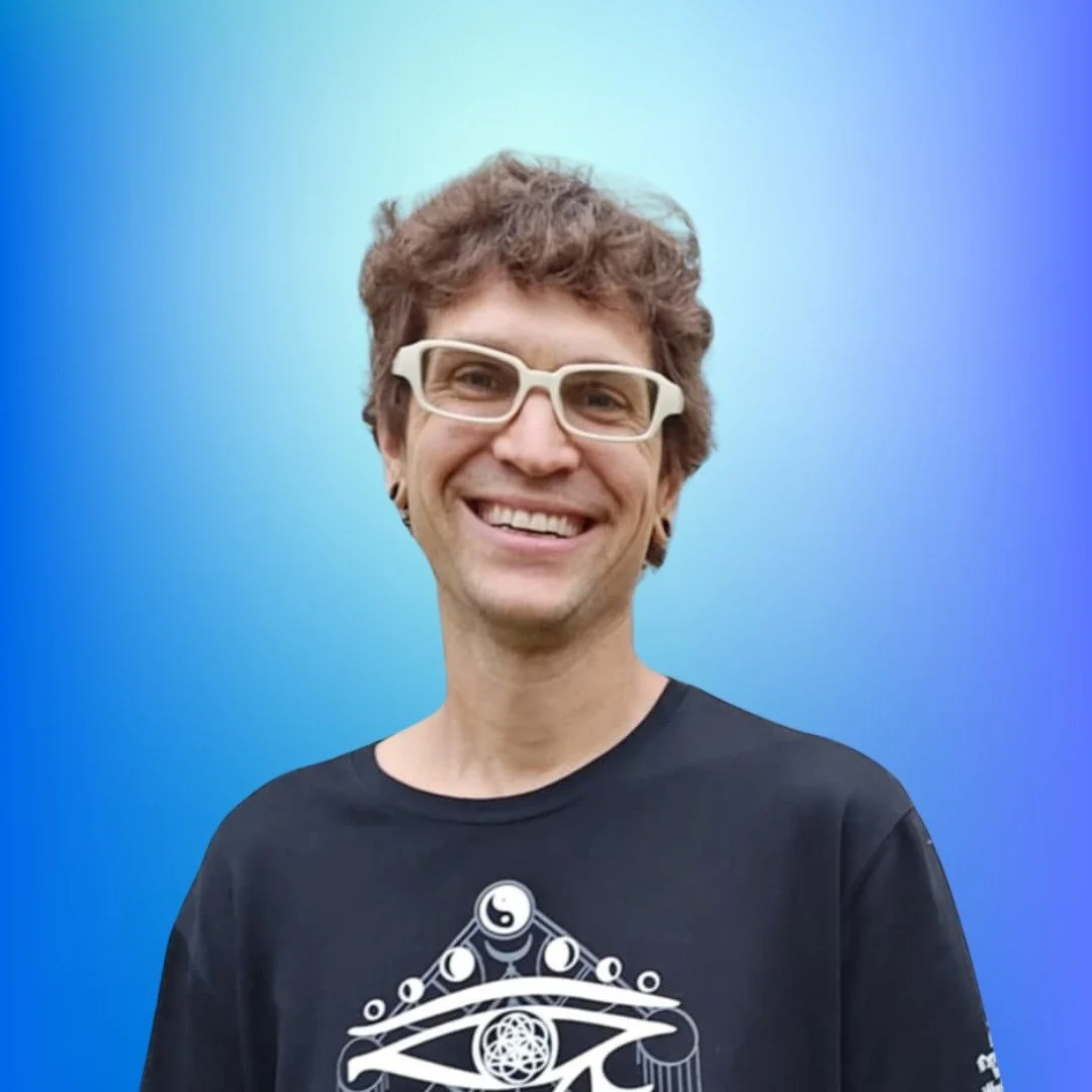 A person with short curly brown hair, white glasses, and a black T-shirt smiling in front of a gradient blue background.