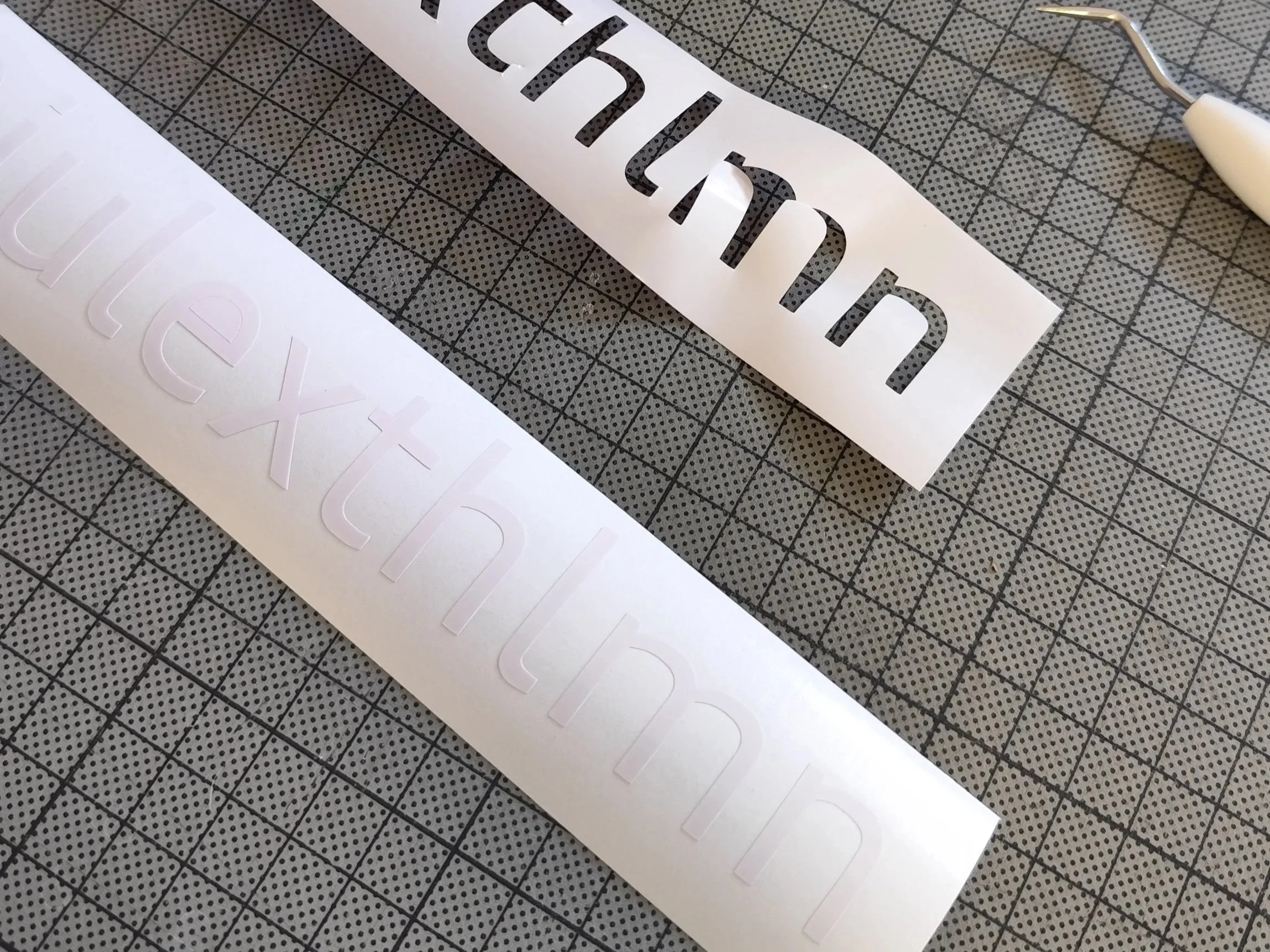 Schriftzüge mit Aufklebern, einer in schwarzen Buchstaben auf weißem Hintergrund und einer in weißen Buchstaben auf transparentem Hintergrund, liegen auf einem Schneide- oder Basteltisch mit Gittermuster.