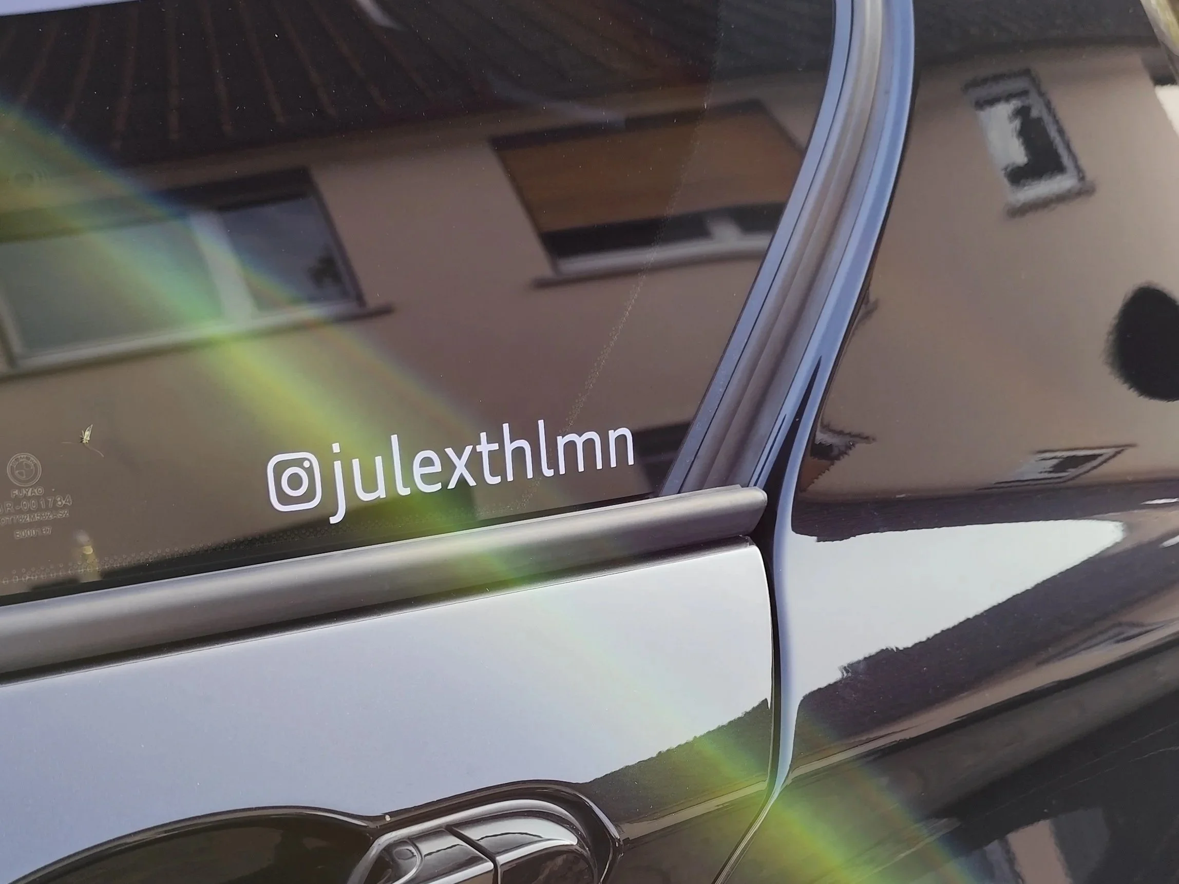 Aufkleber mit dem Instagram-Handle @julexthlmm auf der Heckscheibe eines Autos, im Hintergrund sind Wohnhäuser sichtbar.