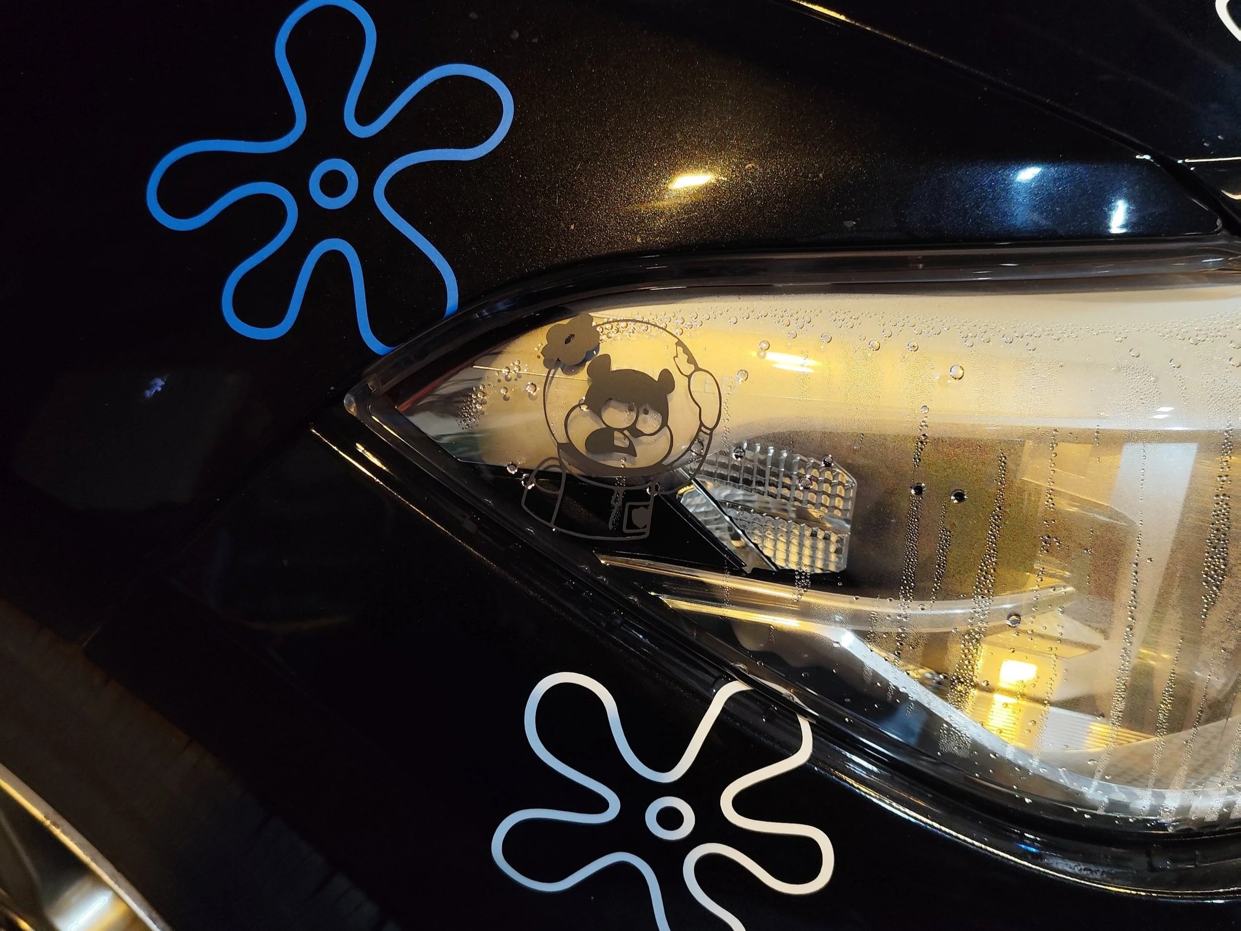 Autolampe mit Wassertröpfchen, auf der eine graue Zeichenzeichnung von Sandy in einem Astronautenanzug ist, zwei blumenförmige Sticker in Blau und Weiß auf schwarzem Fahrzeug