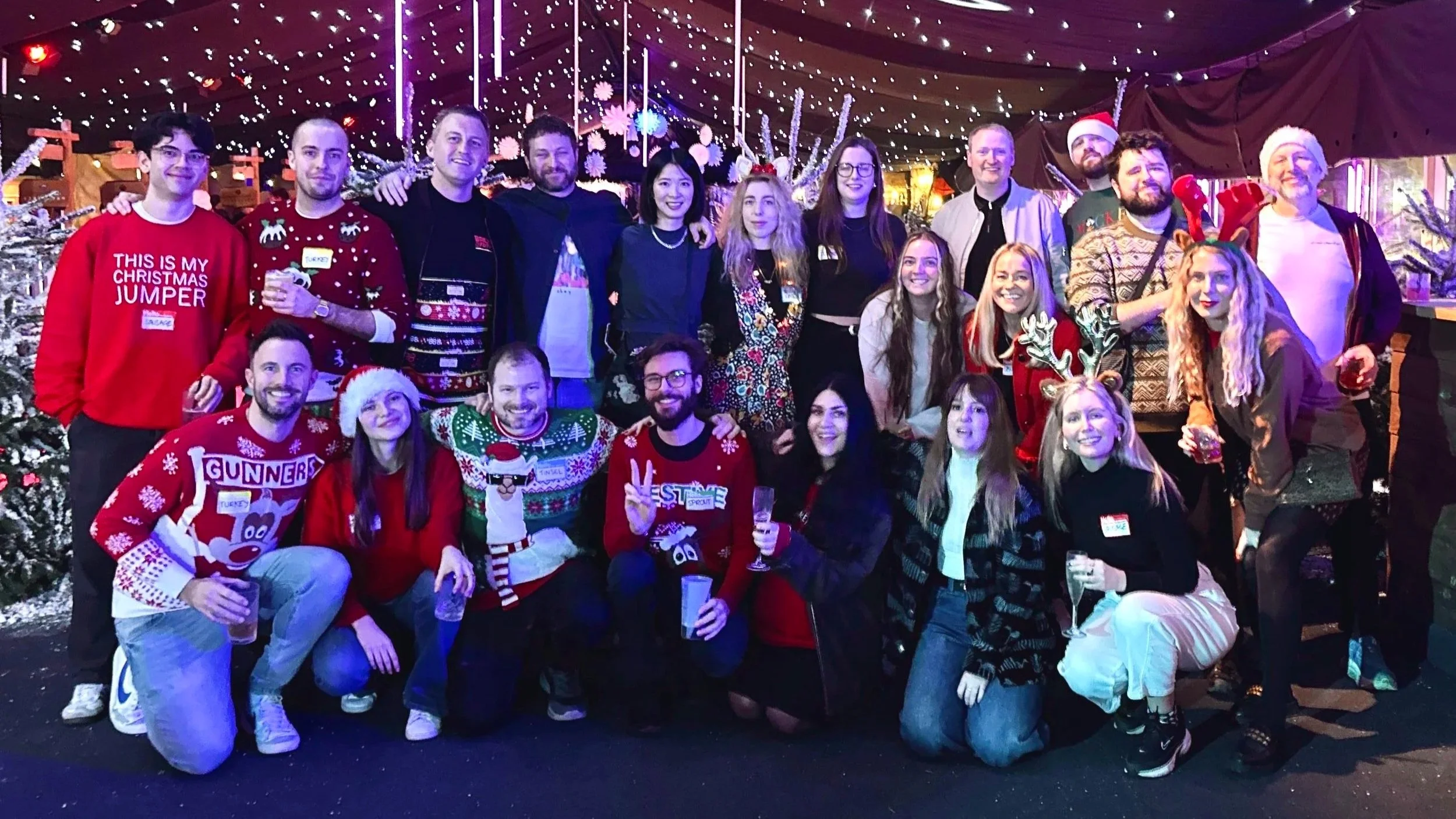Group of 20 people celebrating Christmas, wearing festive sweaters and accessories, in a decorated setting with Christmas trees, string lights, and snow-themed decorations.