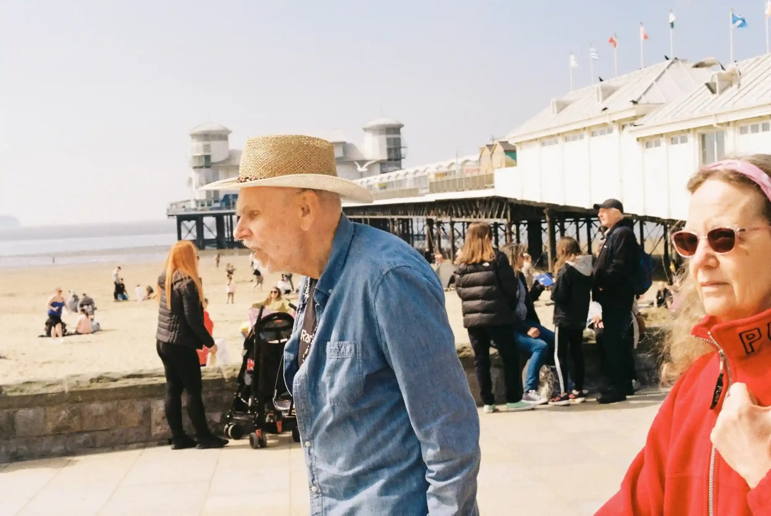 A busy promenade at Weston-super-Mare with people walking in the sun, featuring an older man in a straw hat and denim shirt in the foreground and the white towers of the Grand Pier in the background, by Matthew Morgan.