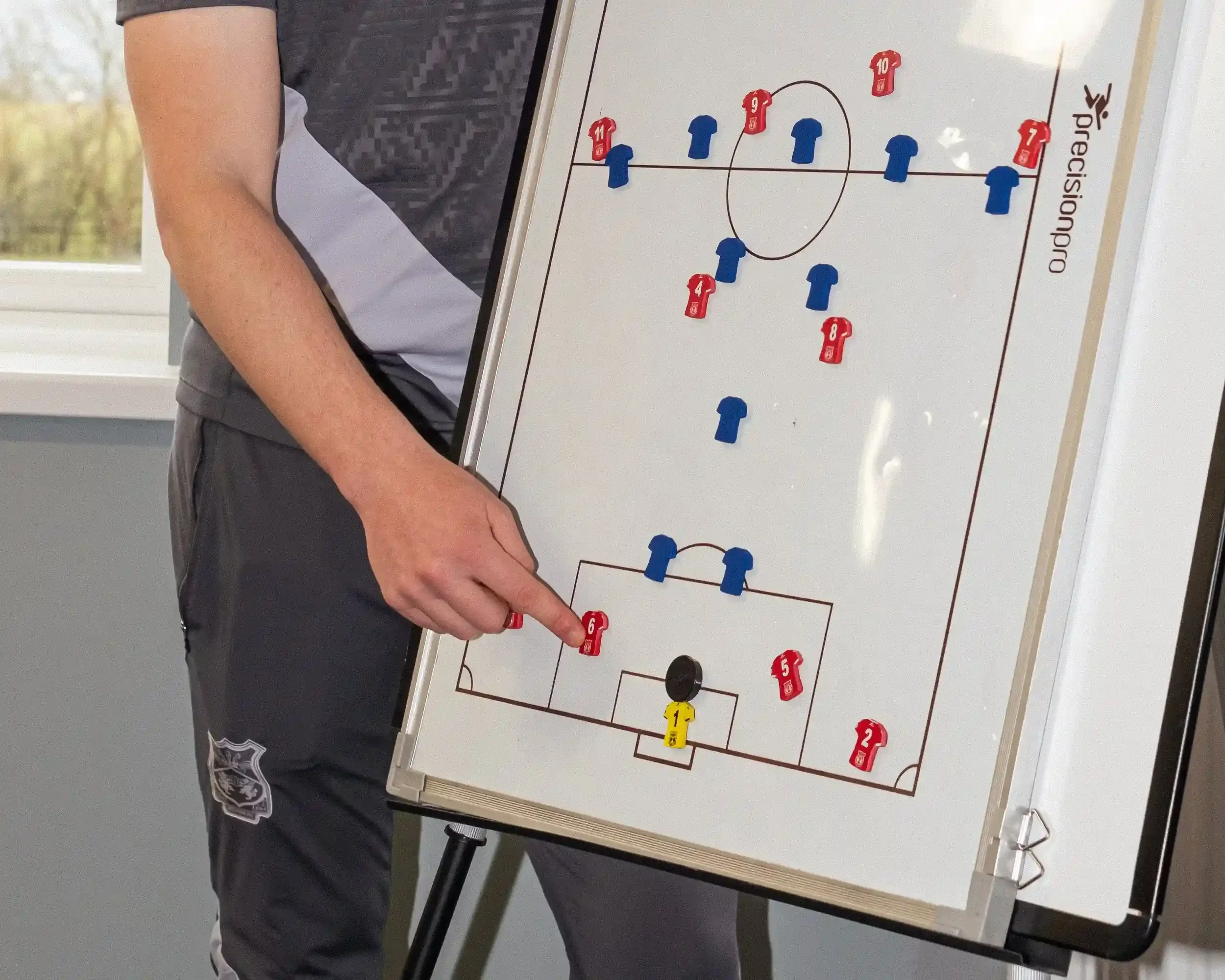 A close-up shot of a football coach’s hand pointing to a tactical whiteboard during a session for Wrexham AFC. The board features a full pitch diagram with various red and blue jersey-shaped tactical magnets from Your Game Your Name. A yellow goalkee