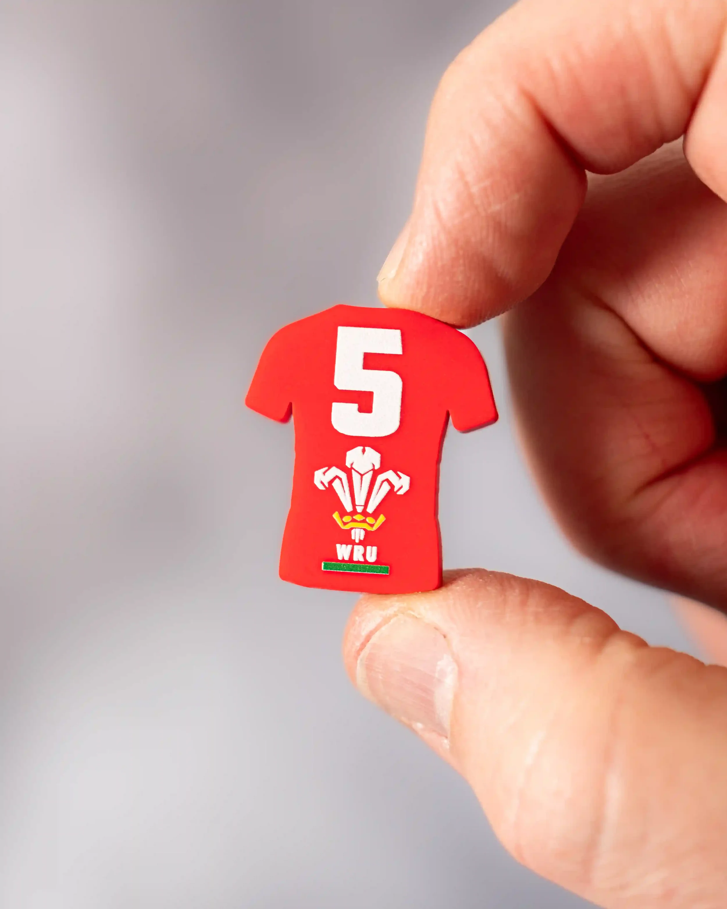A macro close-up of a small, red rubberized magnet shaped like a rugby jersey, held between a person's thumb and finger. The magnet features a large white number '5' above the official Welsh Rugby Union three-feathers logo and 'WRU' text in white. A 