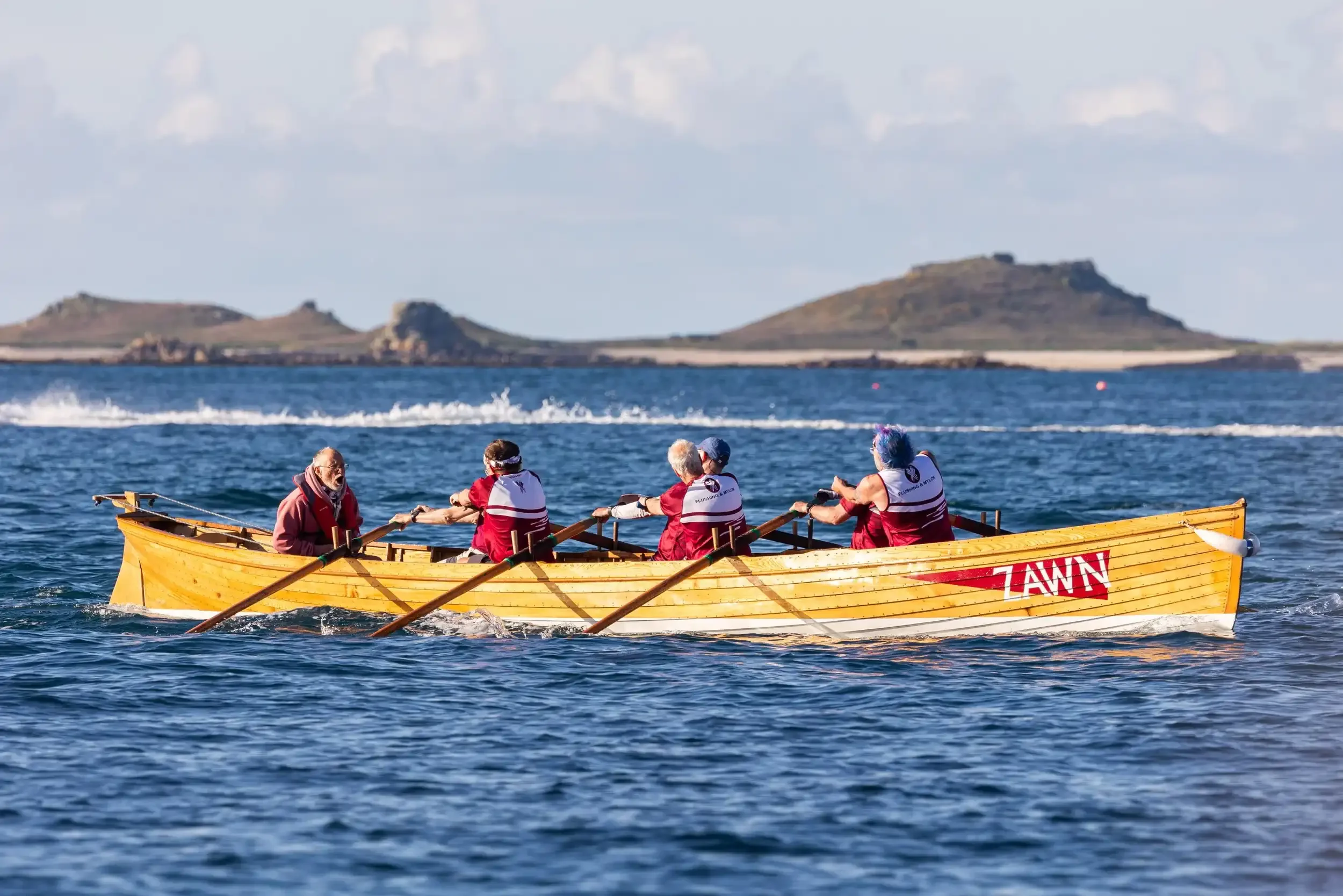 A long shot of a traditional wooden pilot gig boat named 'Zawn' being rowed across choppy blue water by a crew of six. The rowers are wearing maroon and white team shirts, and their wooden oars are extended in a powerful stroke. The coxswain is seate