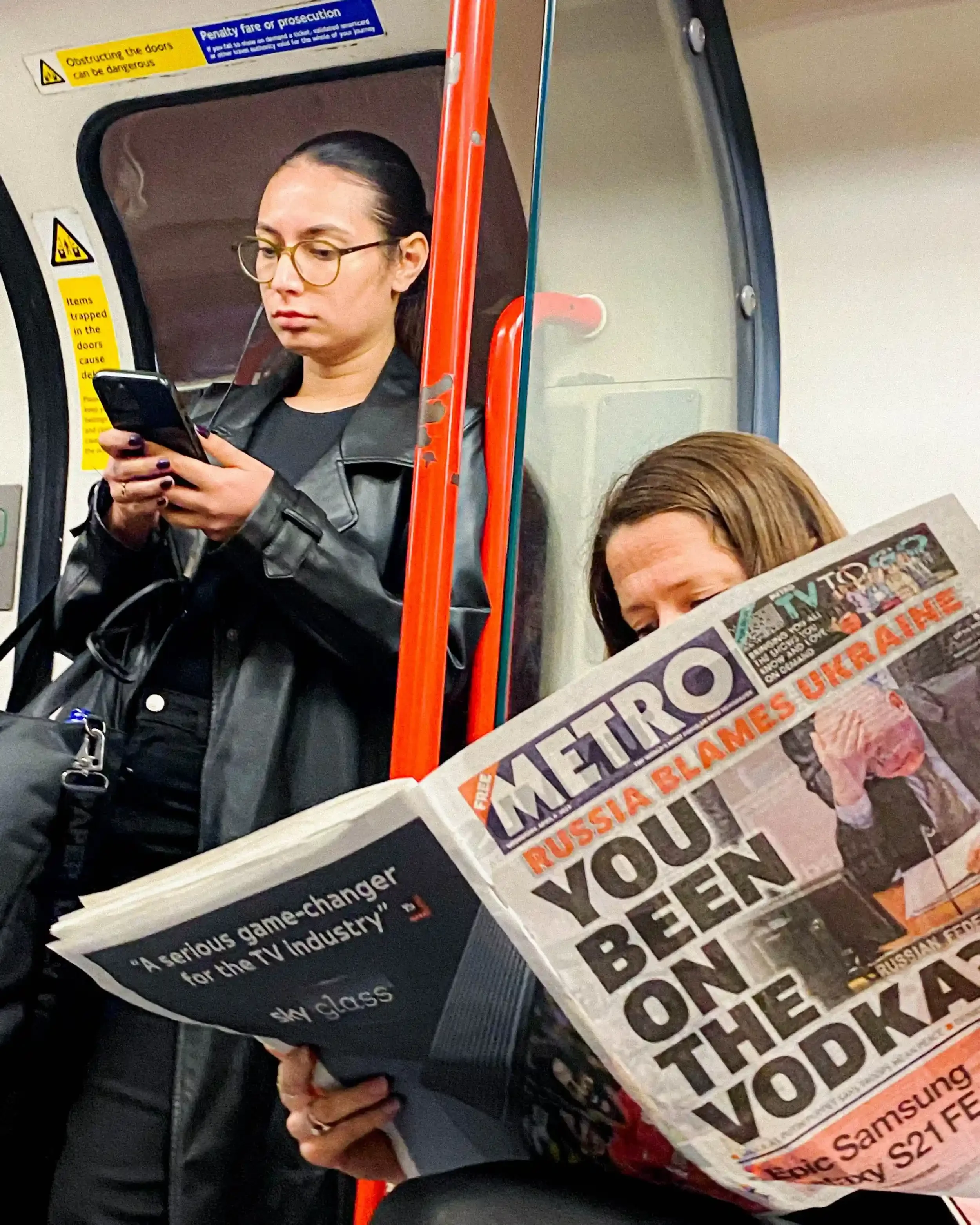 A woman on a London Tube train reading a Metro newspaper with a headline about Russia and Ukraine, while another woman stands behind her using a smartphone, by Matthew Morgan.