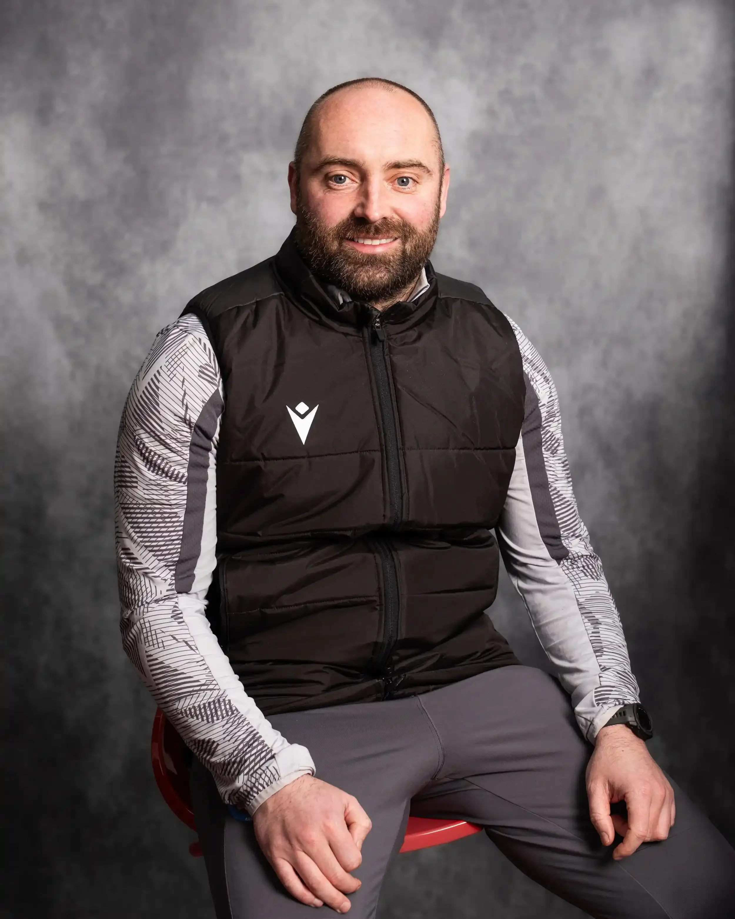 A professional studio portrait of Danny Holmes, an employee at Macron Merseyside. He is a man with a short beard, smiling warmly at the camera while sitting on a red stool. He is wearing a grey Macron training top with patterned sleeves under a black