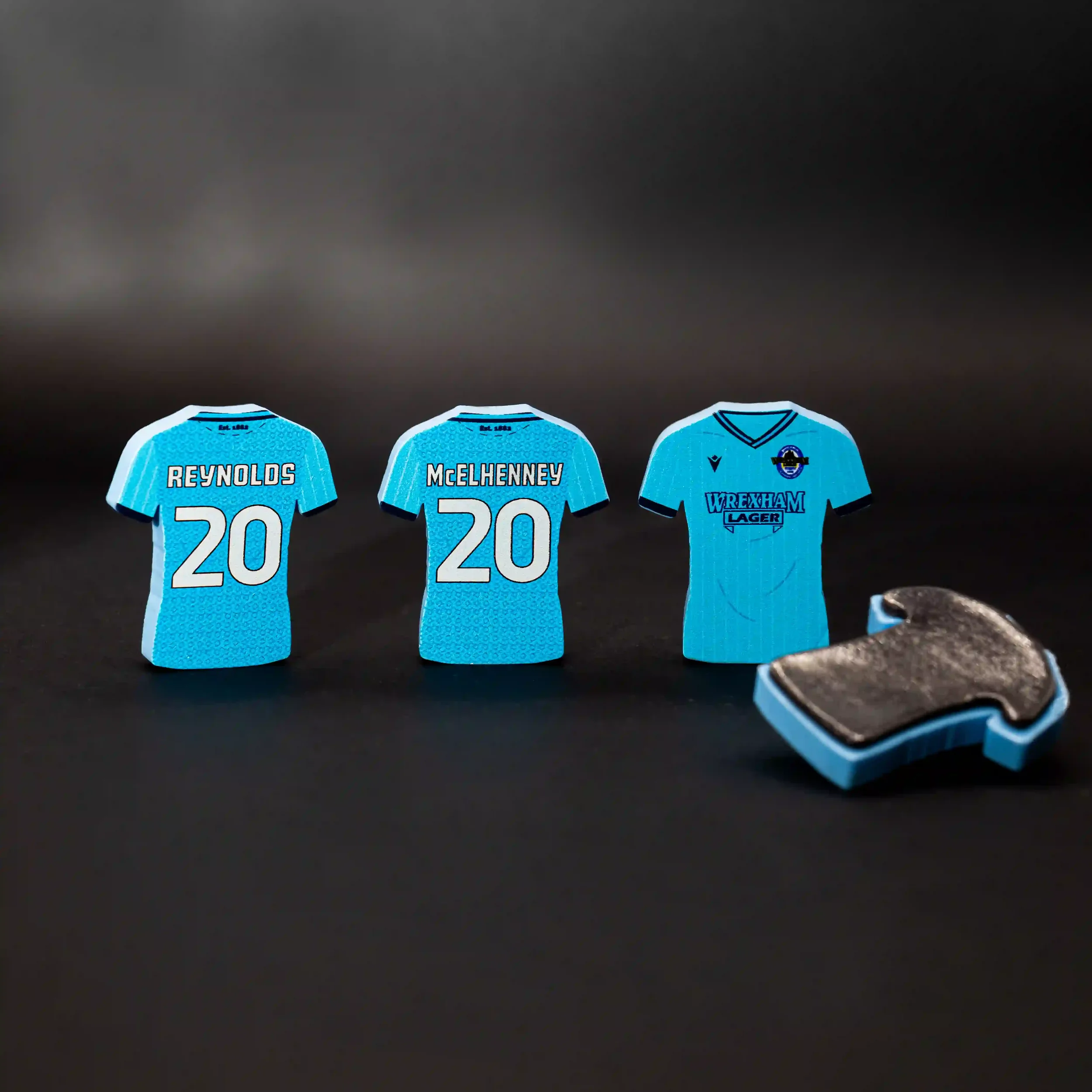 A professional product shot of three light blue shirt-shaped magnets standing against a dark background. On the left, two magnets show the back of a Wrexham AFC jersey with 'REYNOLDS 20' and 'McELHENNEY 20' in white text. The third magnet shows the f