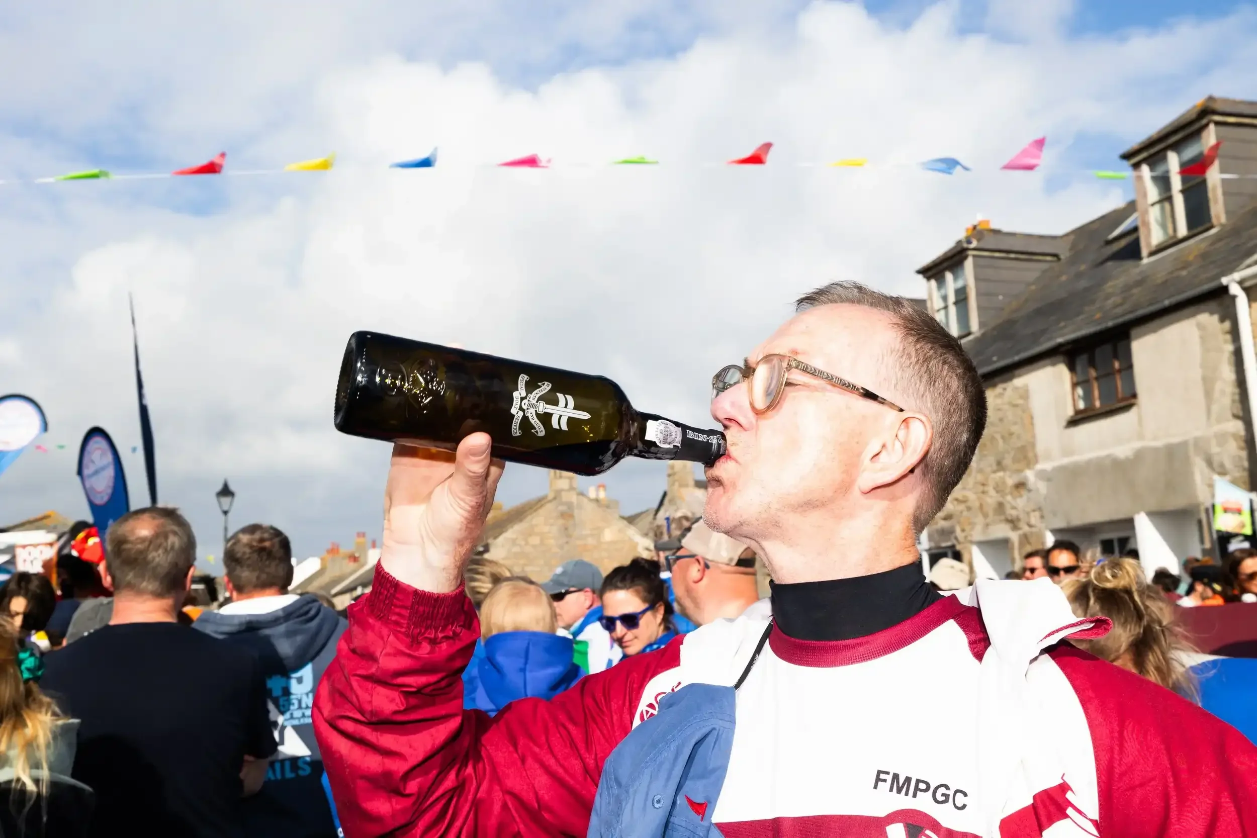 A close-up profile shot of a man with glasses and a maroon and white 'FMPGC' jacket drinking from a dark glass bottle of port. He is standing outdoors in a crowded street during a festival. In the background, colorful bunting hangs against a bright, 