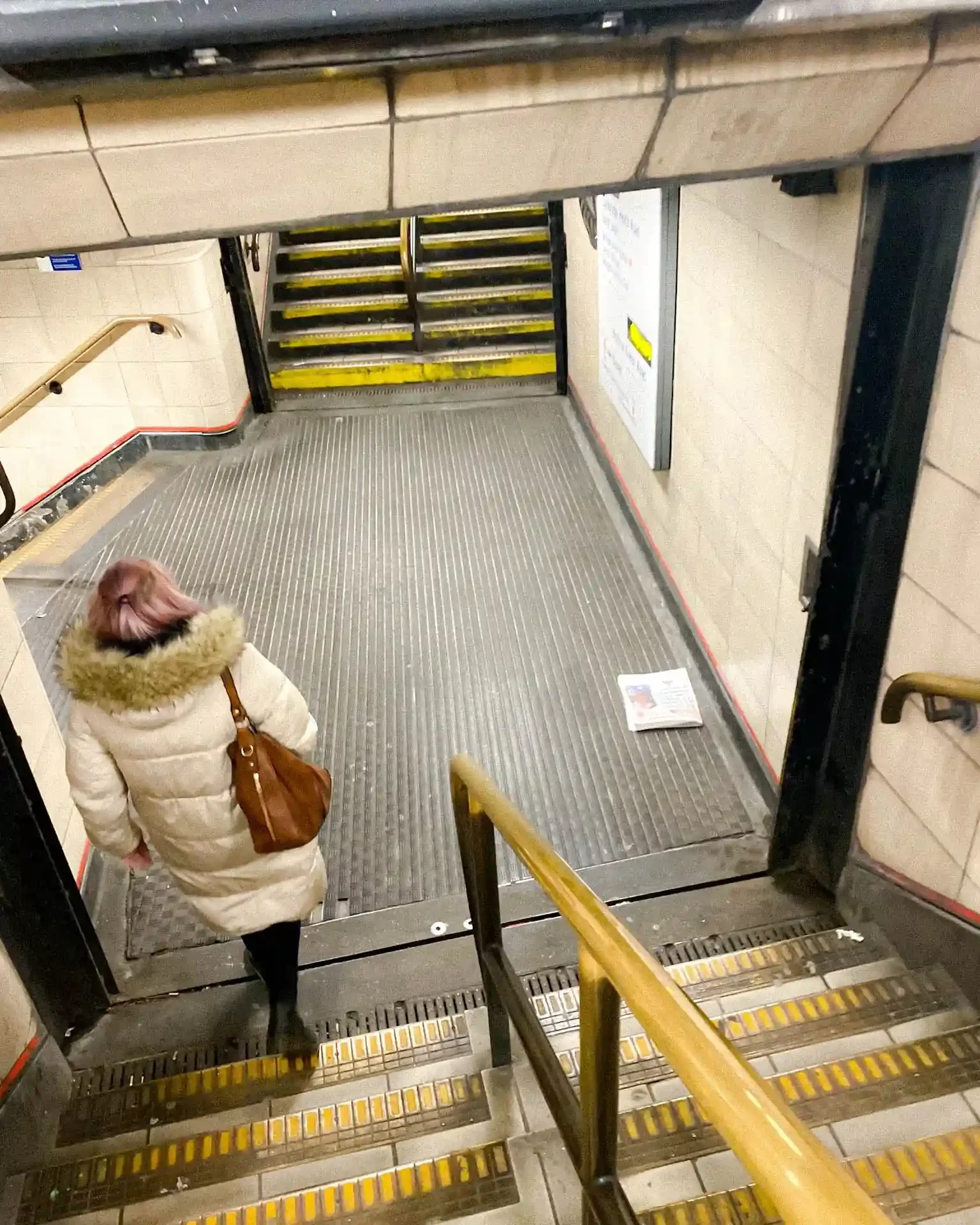 A high-angle shot of a woman with pink hair descending stairs into a London Tube station, passing a discarded newspaper lying on the floor, by Matthew Morgan.