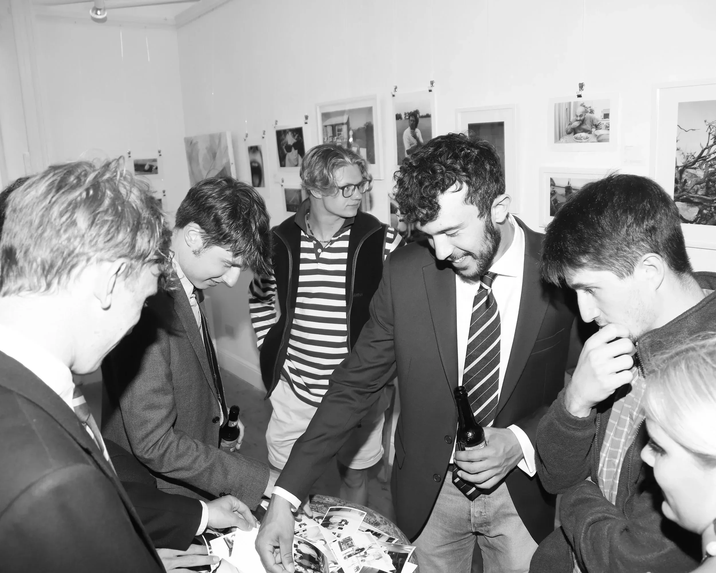 A black-and-white candid photograph of several young people gathered around a rustic metal barrel used as a display table at The Poly gallery in Falmouth. A man in a suit and striped tie is reaching out to touch a pile of small, loose photographic pr