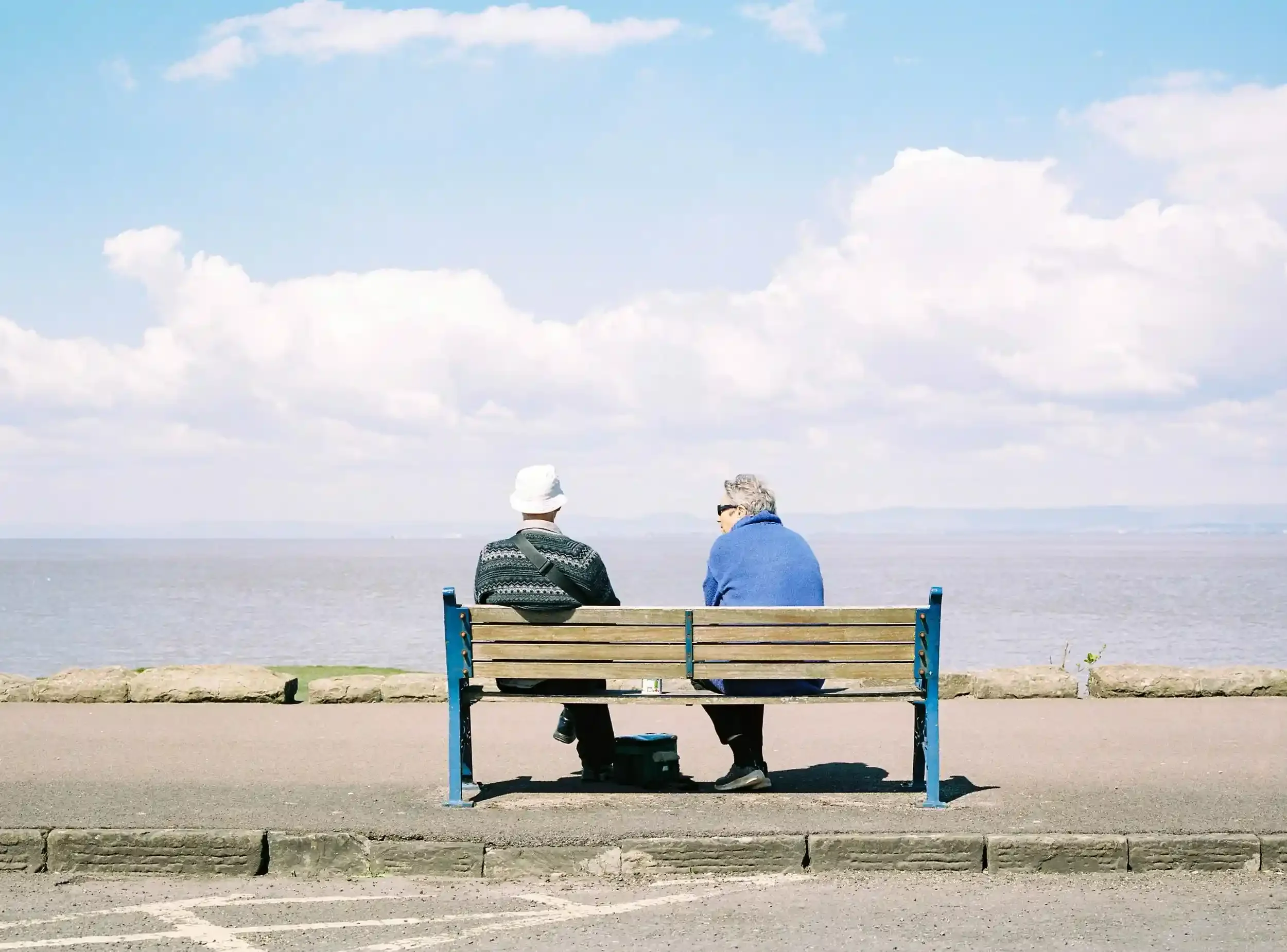 Two people sitting on a blue-framed wooden bench facing a vast, calm sea under a bright blue sky with white clouds in Portishead, by Matthew Morgan.